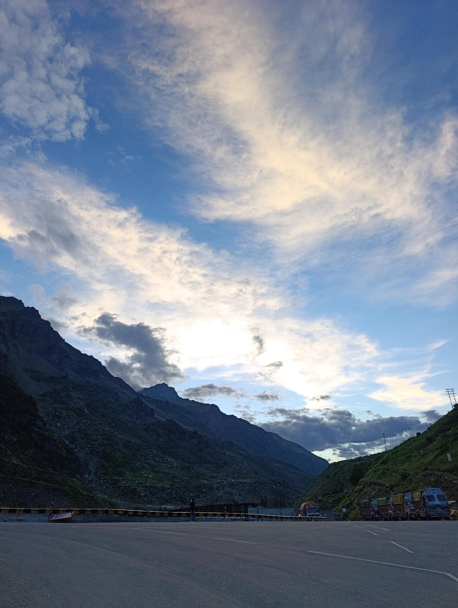 The image shows a wide asphalt road in a mountainous region. On the left, a steep, rocky mountain slope dominates the scene. On the right, another green, sloped mountain rises, with several trucks parked along its base. The trucks appear to be of various sizes and colors, some with loads on their beds. The sky above is a mix of blue with scattered white and pale yellow clouds, suggesting it may be dawn or dusk. In the distance, the mountain range continues, with some peaks shrouded in clouds. There is a yellow and black barrier along the edge of the road. A single person is visible standing near the barrier in the mid-ground. Electrical poles and wires are visible on the right side of the image, against the green slope. The overall impression is a remote, scenic location in India, likely a highway passing through the Himalayas.