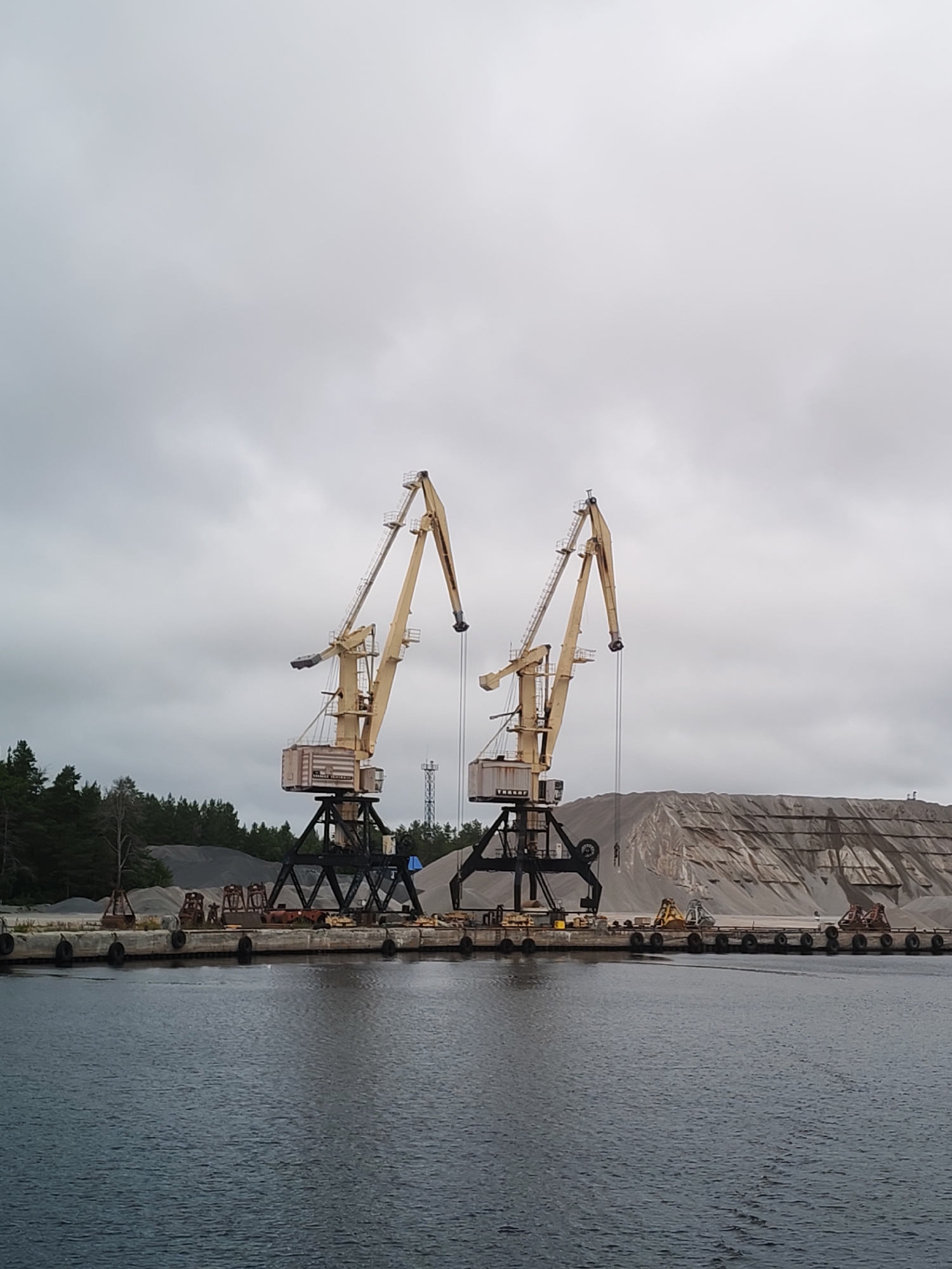 The image depicts a port scene in Shal'skii, Russia, under an overcast sky. Dominating the mid-ground are two large, yellow gantry cranes positioned on a concrete pier. The cranes have intricate metal structures and long, angled jibs. To the right of the cranes, a massive pile of sand or gravel is visible, with terraced layers indicating recent material handling. In the foreground, dark, rippling water stretches across the frame, with a section of the pier visible along the bottom edge. The pier is lined with dark, circular buoys and a few pieces of industrial equipment, including what appear to be smaller cranes or loading machines. A line of dark green trees borders the background on the left, beyond the pier and material piles. A tall, slender tower, possibly a communication tower, is visible in the distance behind the cranes. The overall impression is one of an industrial port engaged in bulk material handling.