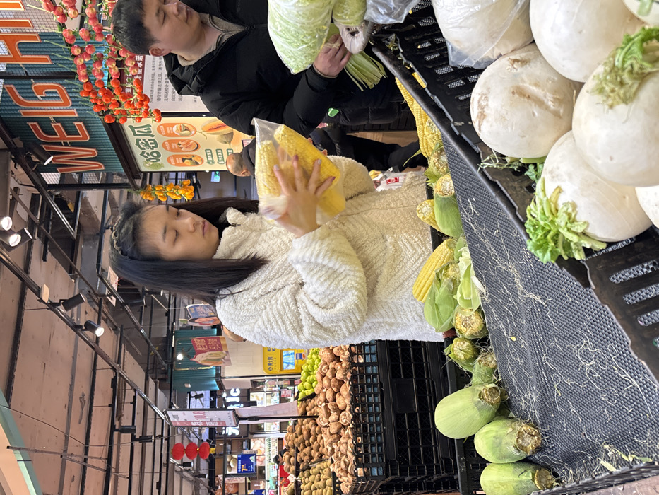 The scene depicts the produce section of a supermarket in Jiangjin, China. A young East Asian female, with dark hair and wearing a cream-colored textured fleece jacket, stands in the foreground, holding and examining a clear plastic bag containing several yellow corn cobs. Her head is tilted downwards, focusing on the corn. To her right, a middle-aged East Asian male in a black puffer jacket is partially visible, also looking down at green leafy vegetables in a plastic bag on an adjacent display.
In the immediate foreground, a black slatted display contains various corn cobs, some partially husked, along with scattered corn silk. Below this, a crate holds large white radishes or turnips with green tops. To the left of the female subject, black crates are filled with brown root vegetables, likely potatoes, and further back, piles of green citrus fruits are visible.
Overhead, industrial-style ceiling with exposed pipes and track lighting is present. A prominent sign in the background displays "WEIGH" in large English letters with corresponding Chinese characters below. Decorative strings of small red and yellow fruit-like objects hang from the display structure. Further in the background, other shoppers are visible, along with additional supermarket shelving, red lanterns, and a red sign displaying "10.0" with Chinese text.