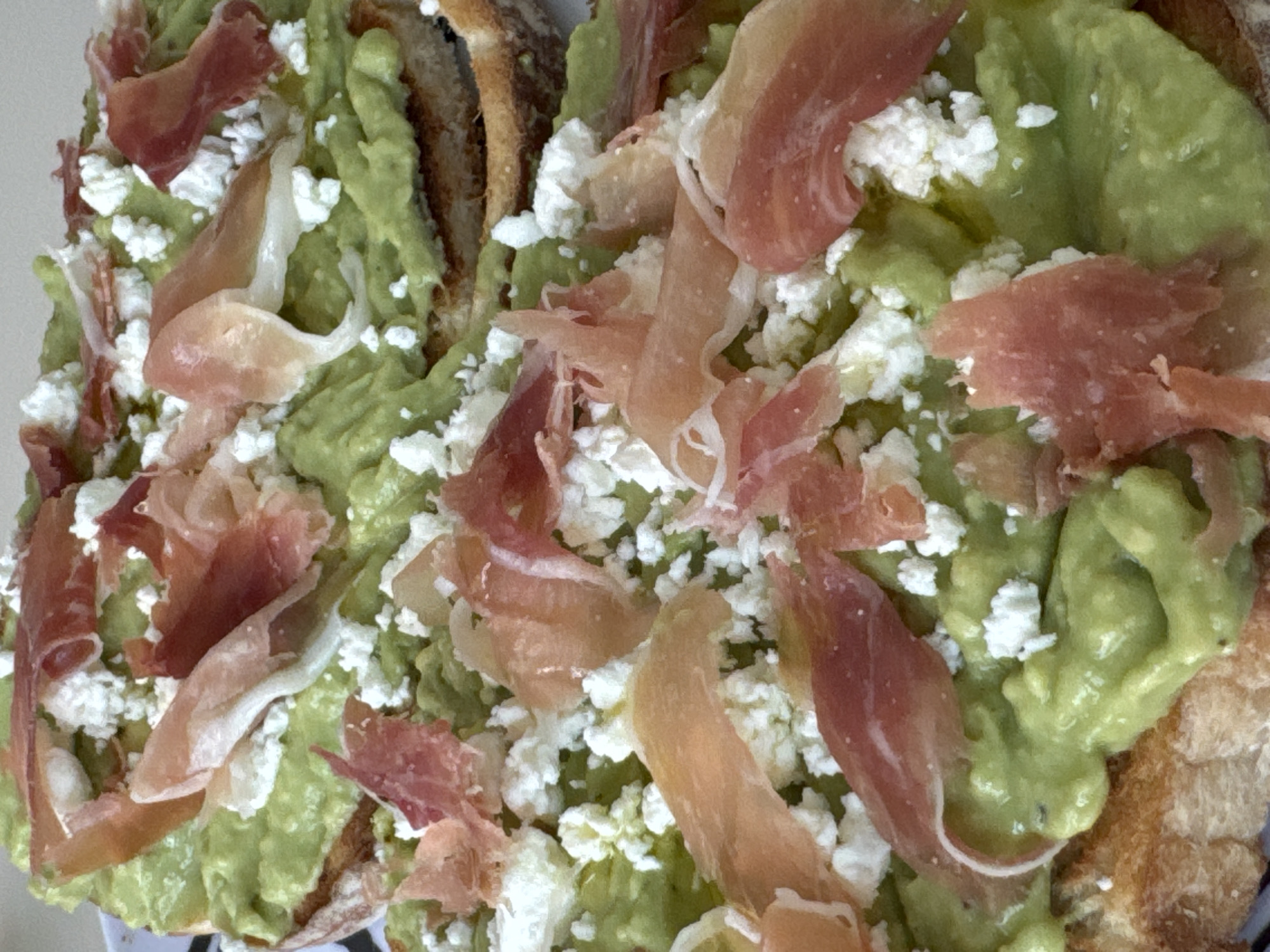 The image shows a close-up of avocado toast topped with crumbled cheese and thinly sliced prosciutto. The bread appears to be toasted and slightly charred. The avocado is mashed and spread across the bread. The crumbled cheese is white, and the prosciutto has a pale pink color with some darker red portions. The overall appearance suggests the food is freshly prepared. The image does not contain location information. Therefore, it cannot be determined whether the image was taken in San Bernardino Tlaxcalancingo, Mexico.