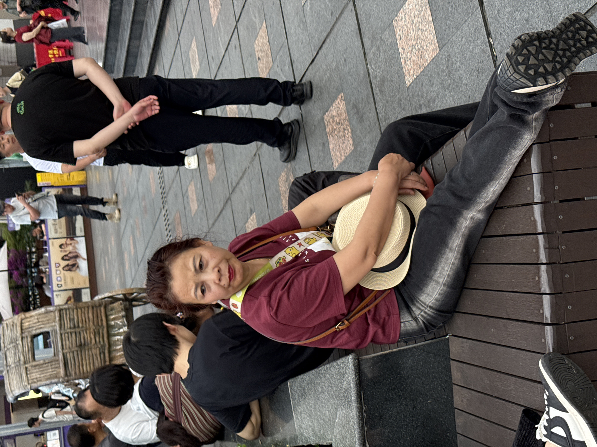 A high-angle shot depicts a woman sitting on a brown wooden bench in an outdoor urban setting. She is wearing a maroon shirt, dark denim pants, and athletic shoes. She holds a straw hat in her lap. Other individuals are present; one person is directly beside her and seems to be bowing their head and back. Still other individuals are walking in the background. The location is Yuzhong District, China.