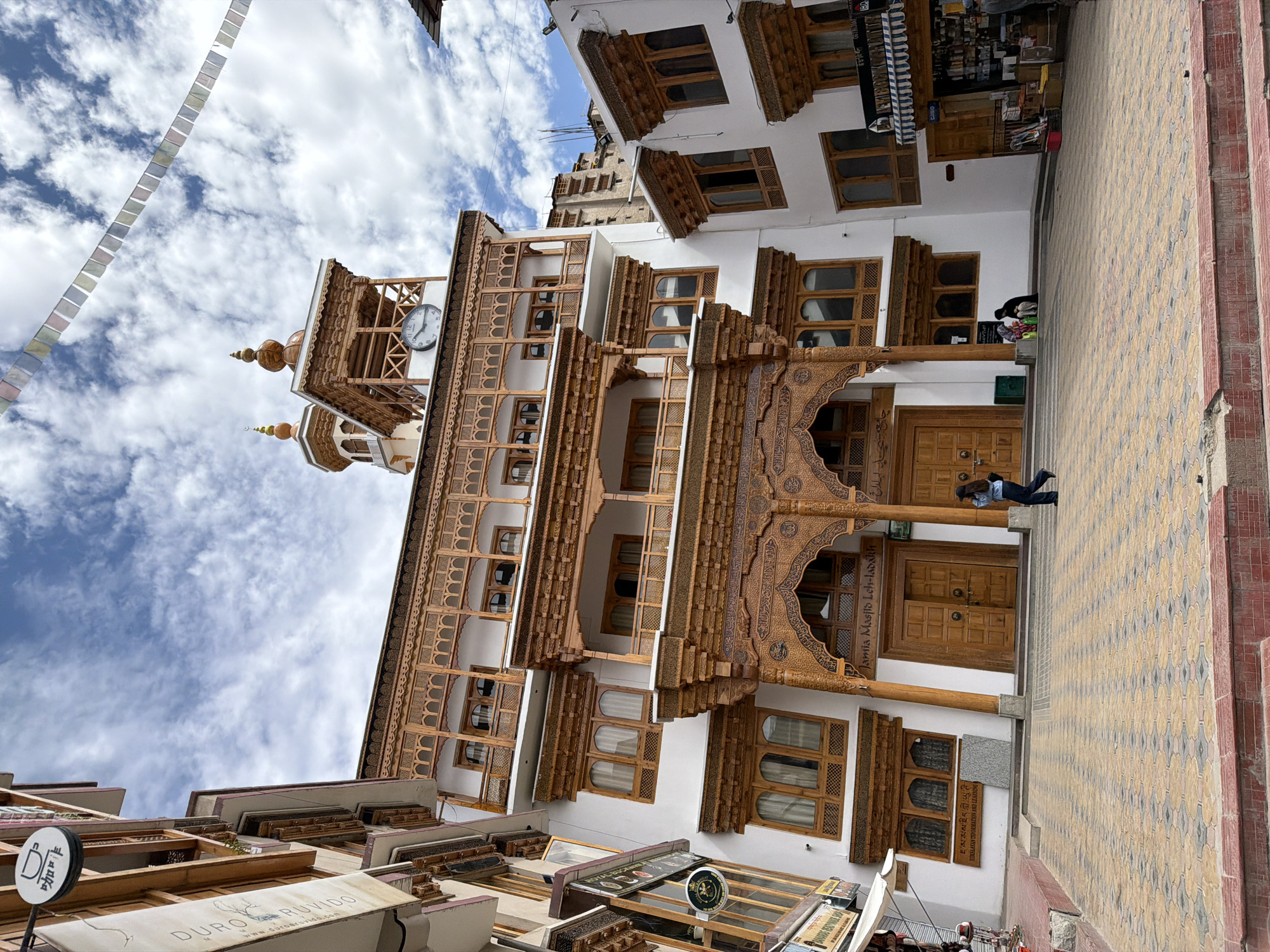 The image shows the Jama Masjid mosque in Leh, India. The mosque is a white building with traditional Ladakhi architecture, featuring wooden carvings and balconies. The mosque has a central courtyard paved with colorful tiles. A person in a blue shirt and black pants is walking across the courtyard. In the background, prayer flags are strung across the sky, and there is a clock tower on top of the mosque. The sky is cloudy, with patches of blue. There are also shops on either side of the mosque.