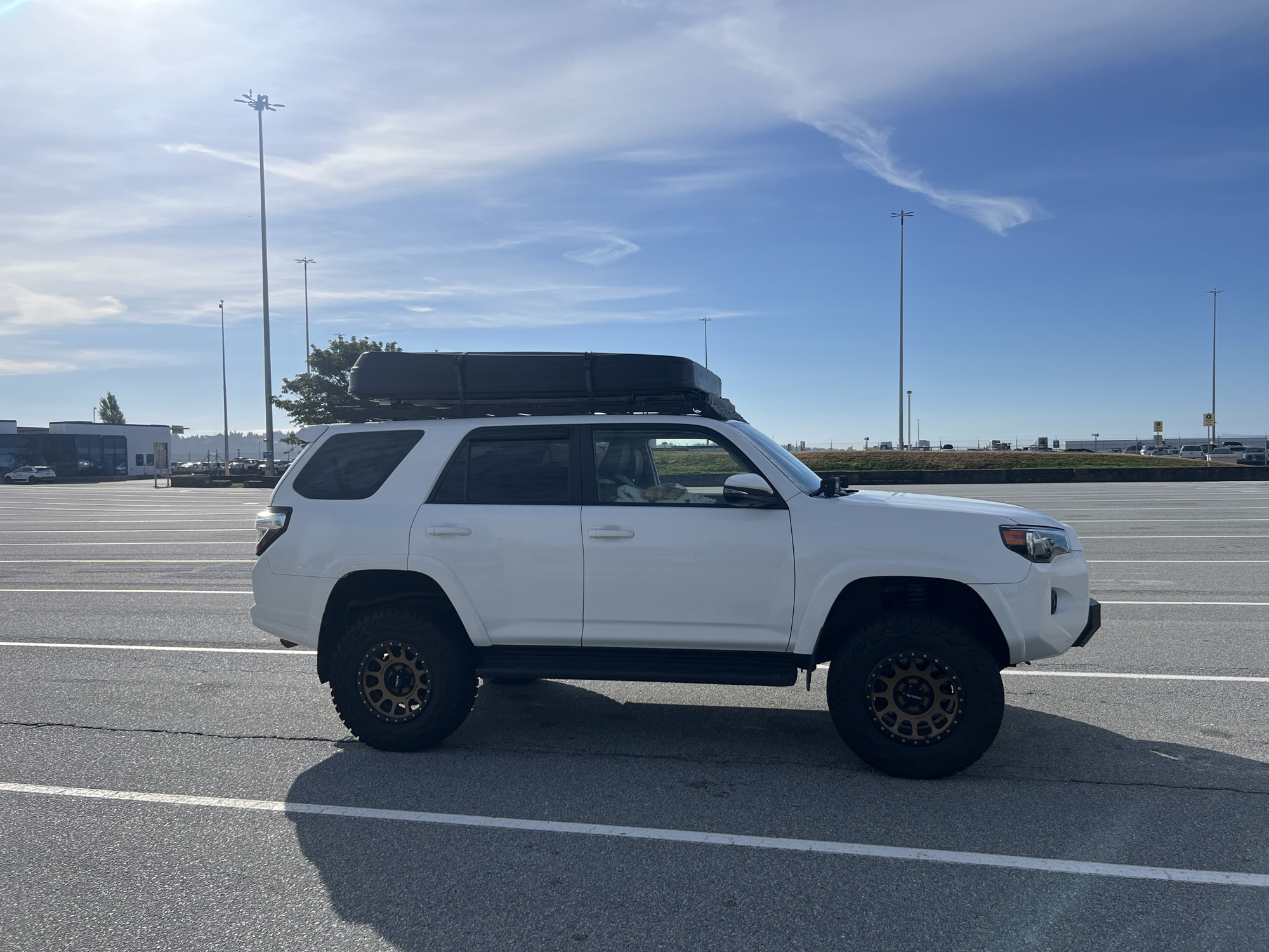 A white Toyota 4Runner SUV, equipped with a black rooftop cargo box and aftermarket bronze-colored wheels, is parked in a large, empty asphalt lot. The vehicle is positioned in the center of the frame, facing right, with its side visible. The ground is marked with white lines, indicating parking stalls or lanes. In the background, several tall lampposts stand under a bright blue sky with scattered white clouds. Distant buildings and trees are also visible on the horizon. The scene appears to be an outdoor parking area, possibly at an airport or a large commercial complex in the city of Delta, Canada, given the presence of light poles typical of such locations. No people are explicitly visible in a distinct interaction, though a person can be partially seen inside the driver's side window of the SUV.