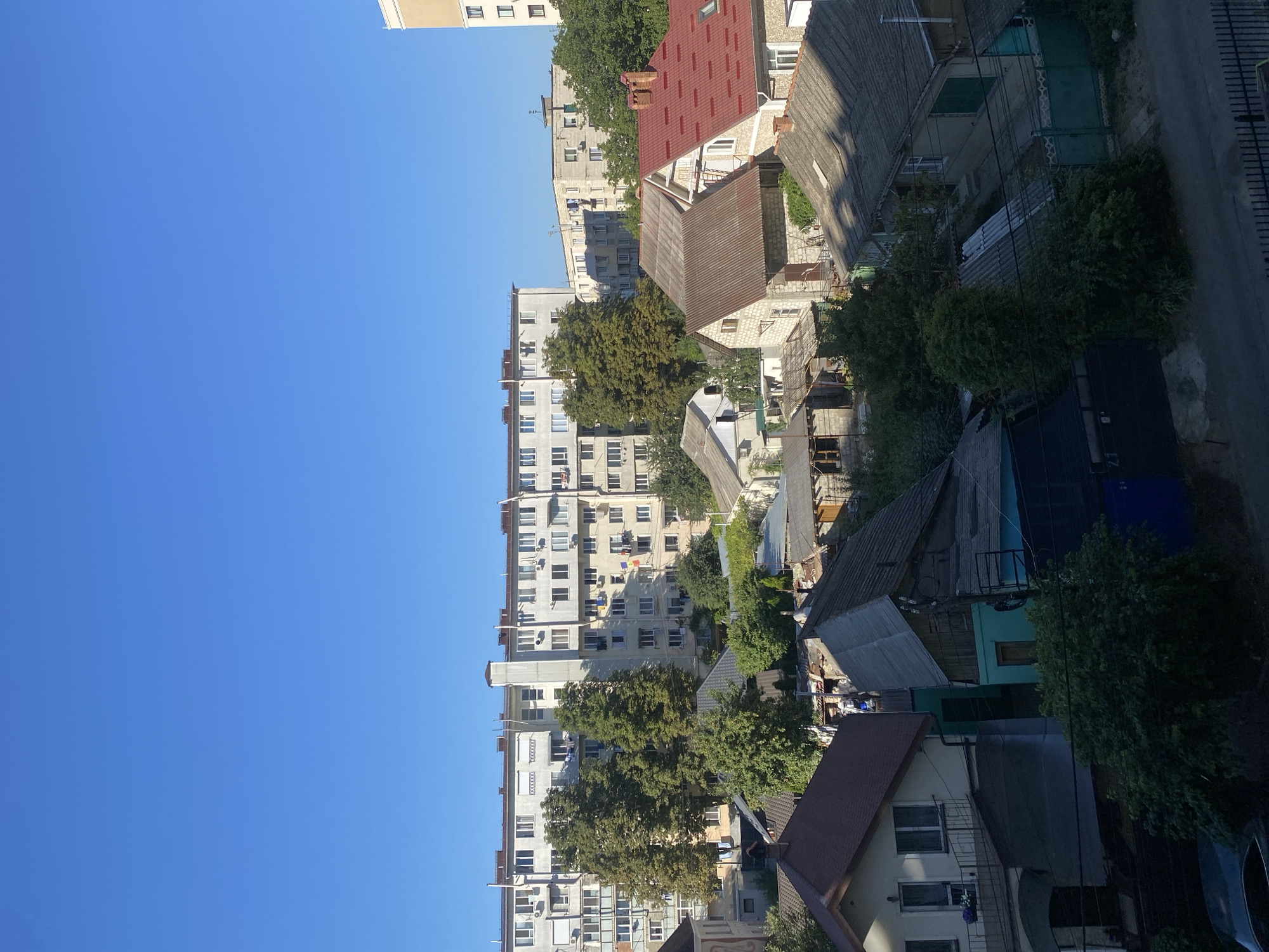 The image shows a view of a residential area in Chișinău, Moldova, on a clear day. In the background, there is a large, light-colored apartment building with many windows and air conditioning units. In front of the apartment building, there are several smaller houses with various roofing materials, including red tiles and corrugated metal. Trees are scattered throughout the scene, some with full green foliage. The foreground shows more houses and a street with a dark-colored car parked partially visible. The overall impression is one of a typical urban residential neighborhood.
