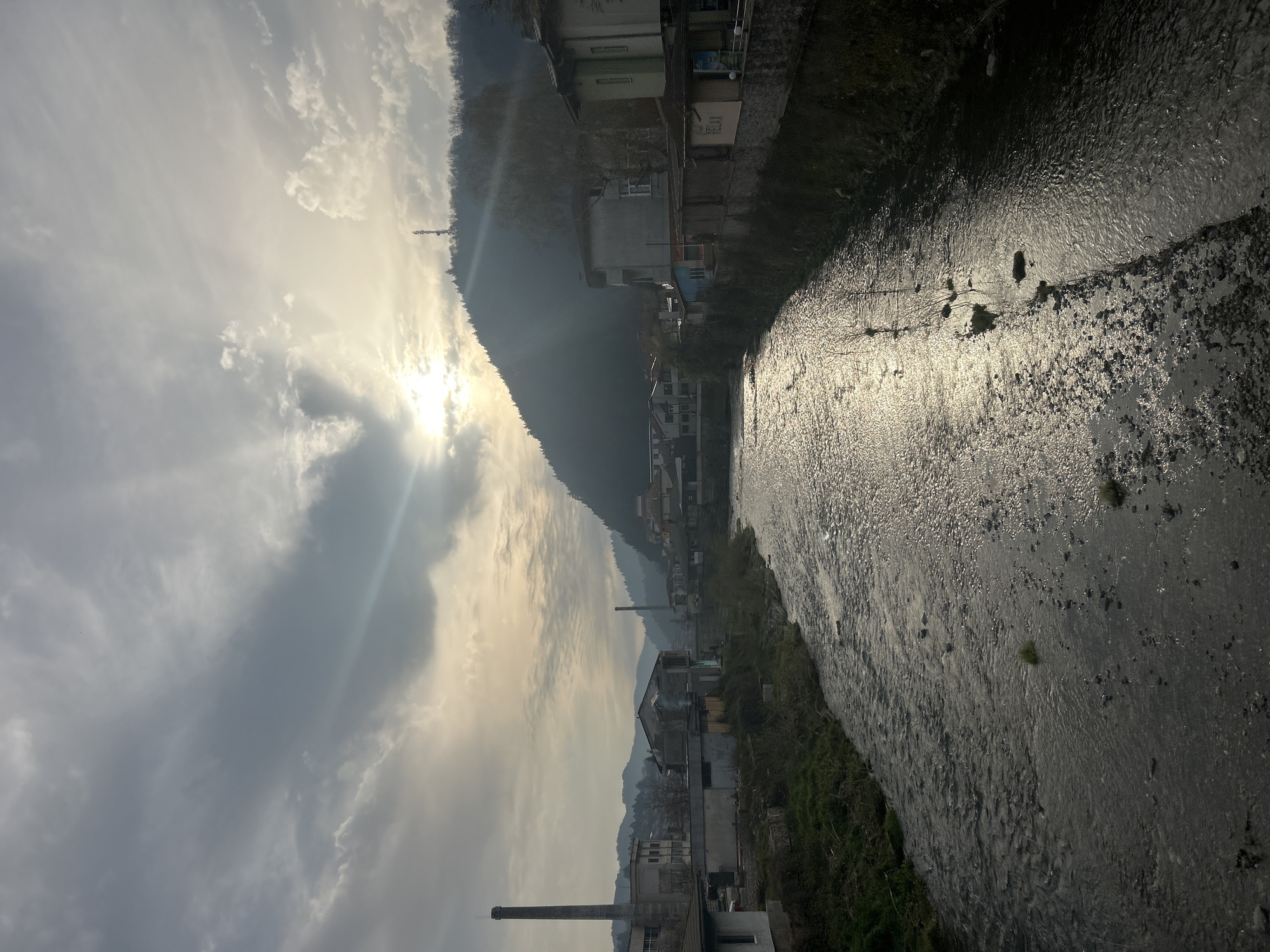 The image captures a view in Zlatograd, Bulgaria. A flowing river bisects the scene, reflecting sunlight. Along the riverbank, buildings line the edge, interspersed with vegetation. A mountain rises in the background, partially obscured by mist or cloud cover. Sunlight filters through the clouds, casting rays across the sky. A tall industrial chimney stands prominently among the buildings in the foreground.