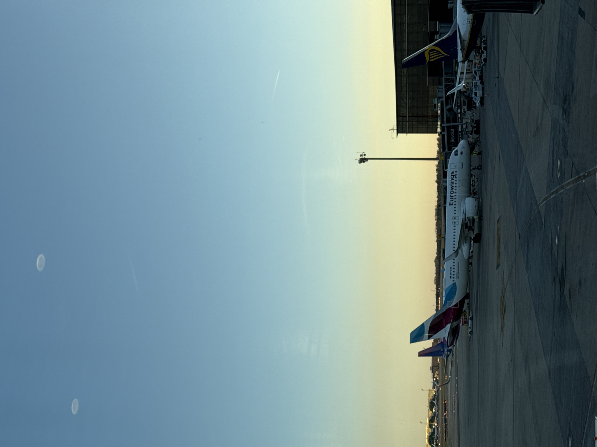 The image depicts an airport scene at El Prat de Llobregat, Spain. Several commercial airplanes are parked at the terminal, including a Eurowings aircraft and a Ryanair plane. A tall light pole stands between the terminal building and the aircraft. The sky transitions from blue to a yellow-orange hue, indicating either sunrise or sunset. Two faint circular shapes are visible in the upper-left sky. The ground is concrete.