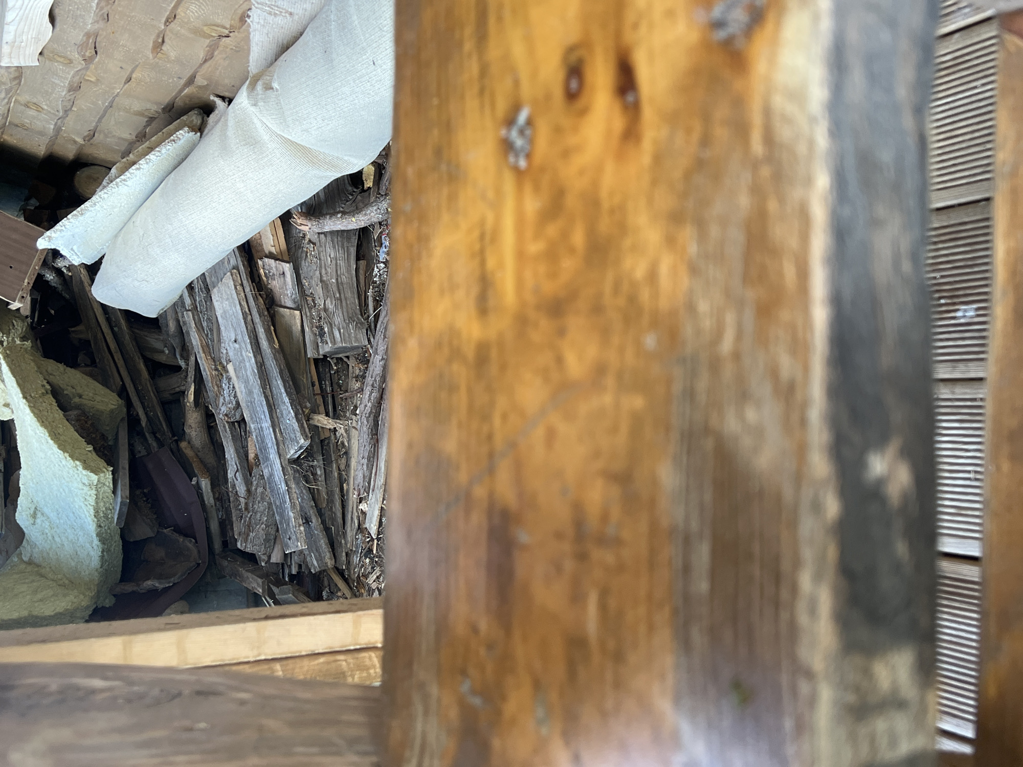 The image depicts a cluttered interior space, likely a shed or storage area, with building materials and debris. In the foreground, a smooth, wooden surface, possibly a table or bench, is in sharp focus. Behind it, the background is a jumble of materials including insulation, wood planks, and what appears to be a rolled-up fabric or tarp. The lighting suggests an indoor environment, possibly with natural light filtering in. The location is identified as Norviliškės, Lithuania.
