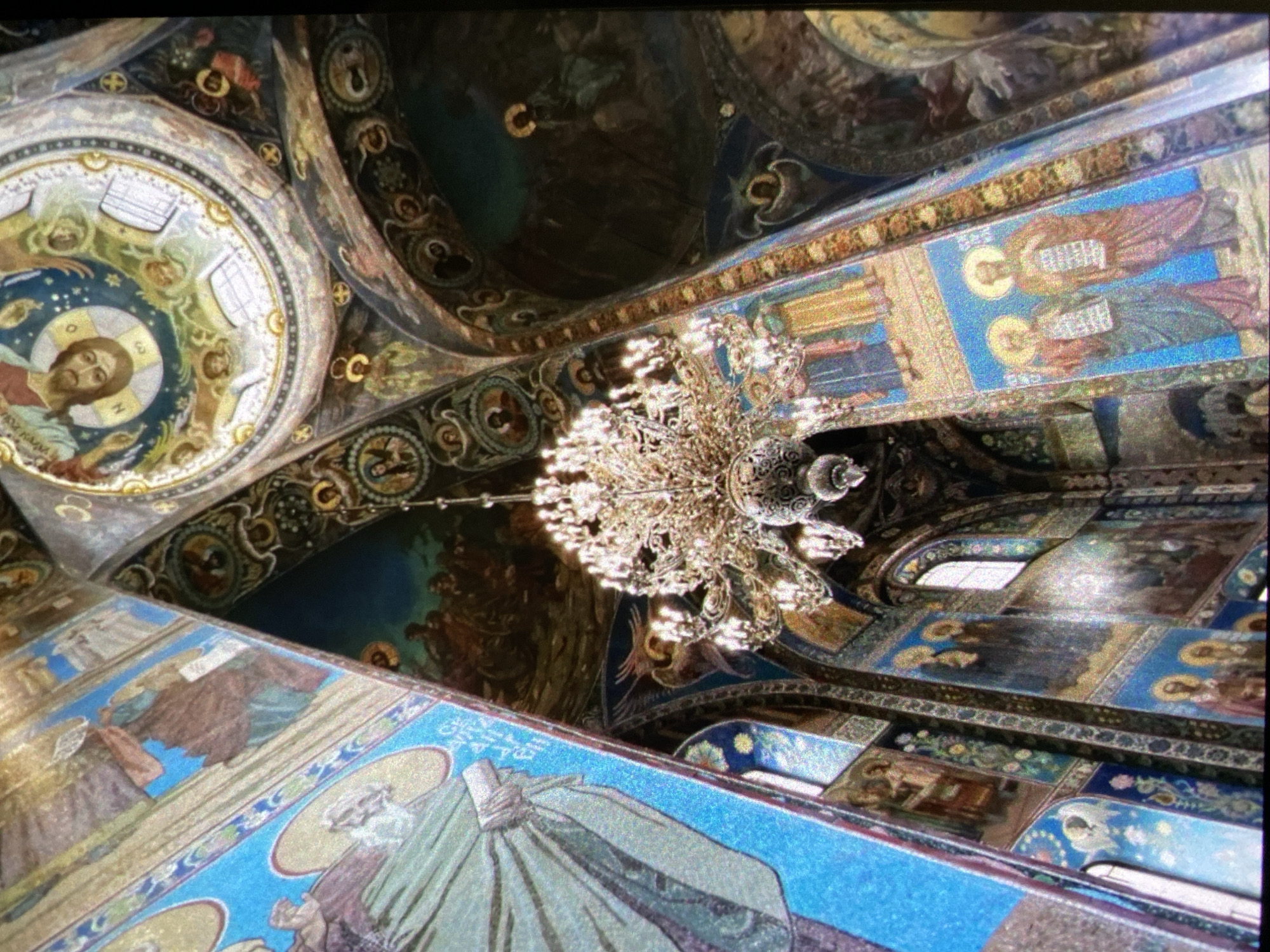 The image captures the interior of the Church of the Savior on Spilled Blood in Saint Petersburg, Russia. The scene is dominated by ornate mosaic artwork covering the walls and ceilings. A large, intricate chandelier hangs from the ceiling. The mosaics depict religious figures. The environment suggests a grand, historic religious setting.