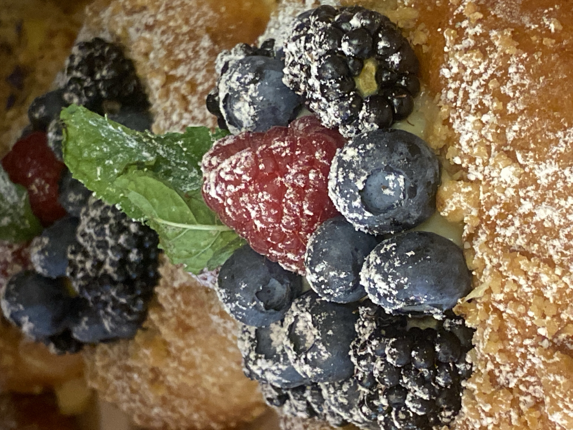 A close-up, overhead shot showcases a dessert generously dusted with powdered sugar. The dessert appears to be a pastry, possibly a croissant or danish, with a flaky, golden-brown texture. It is adorned with a vibrant assortment of fresh berries, including plump blueberries and raspberries, along with dark blackberries. A sprig of fresh mint adds a touch of green to the composition. The powdered sugar creates a light, snowy effect across the pastry and the fruit, suggesting a sweet and indulgent treat. The lighting is bright and even, highlighting the textures and colors of the dessert. The background is blurred, focusing all attention on the delectable pastry. There is no visible text. The context of Warsaw, Poland, is provided but does not offer specific visual cues within the image itself.