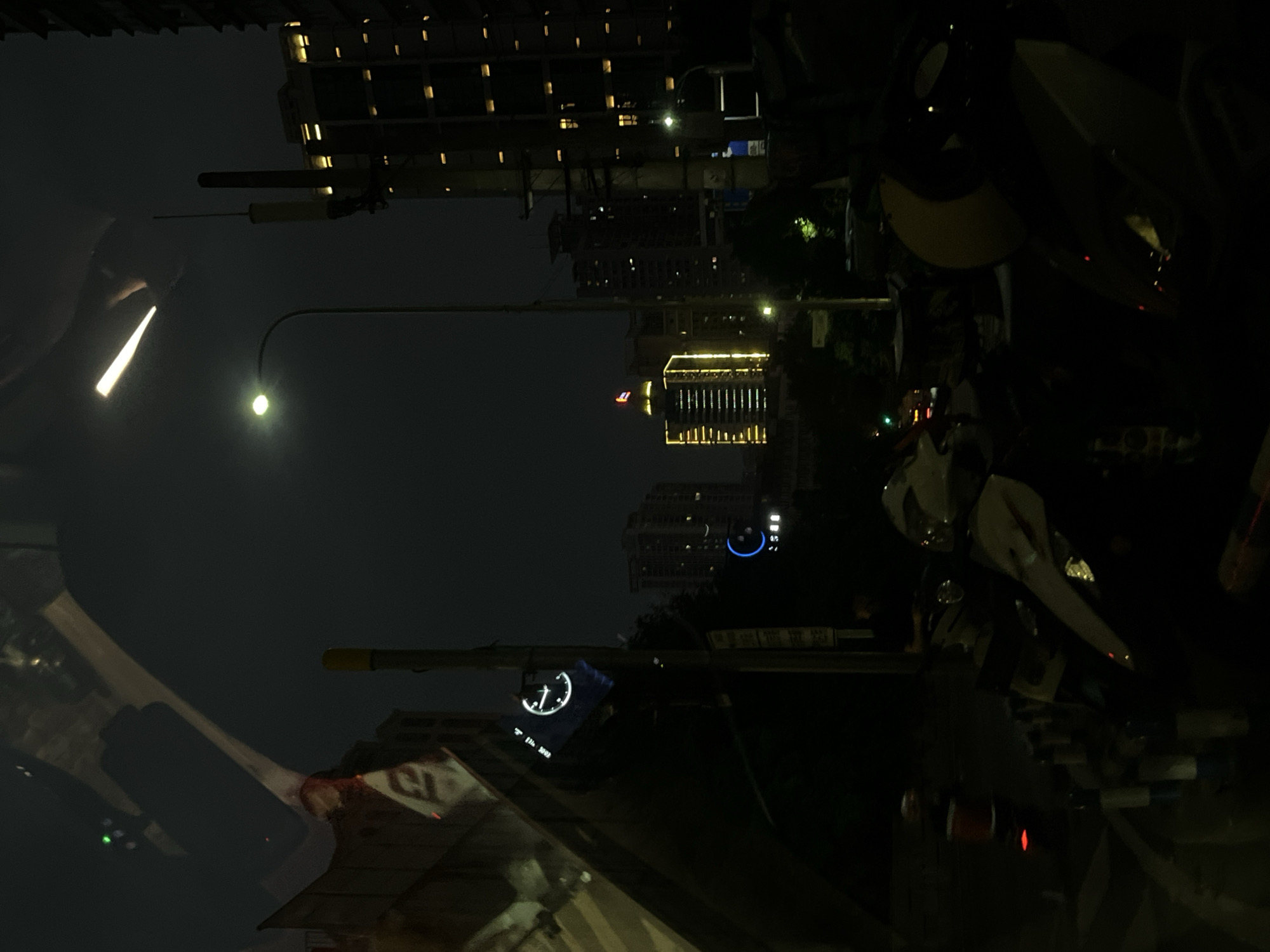 Captured at night in 龙溪街道, China, the image shows a cityscape illuminated by artificial lights. Tall buildings with multiple lit windows dominate the backdrop. A street lamp casts a bright glow on the scene. The foreground features objects suggesting a roadside perspective, including a partially visible vehicle and other urban elements.