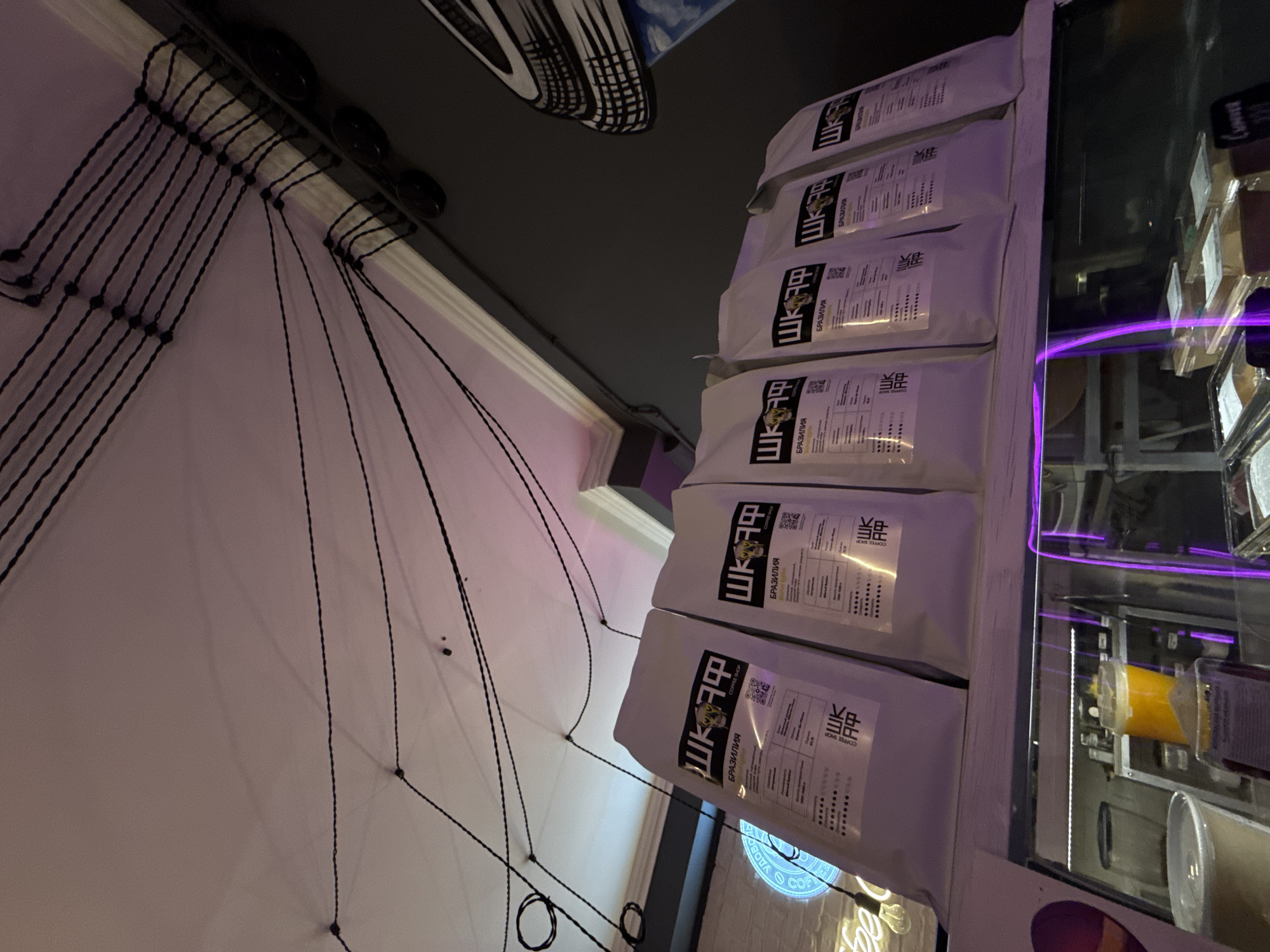 The image shows a display of coffee bags in a shop. The bags are white with black and yellow text and graphics. They are arranged in a row on a shelf. In the background, there are strings hanging from the ceiling and a neon sign. Below the shelf, a refrigerated display case contains various items, including drinks and what appear to be food items. The overall impression is of a modern, possibly trendy, coffee shop. The specific location in Moscow, Russia, cannot be determined from the image alone.