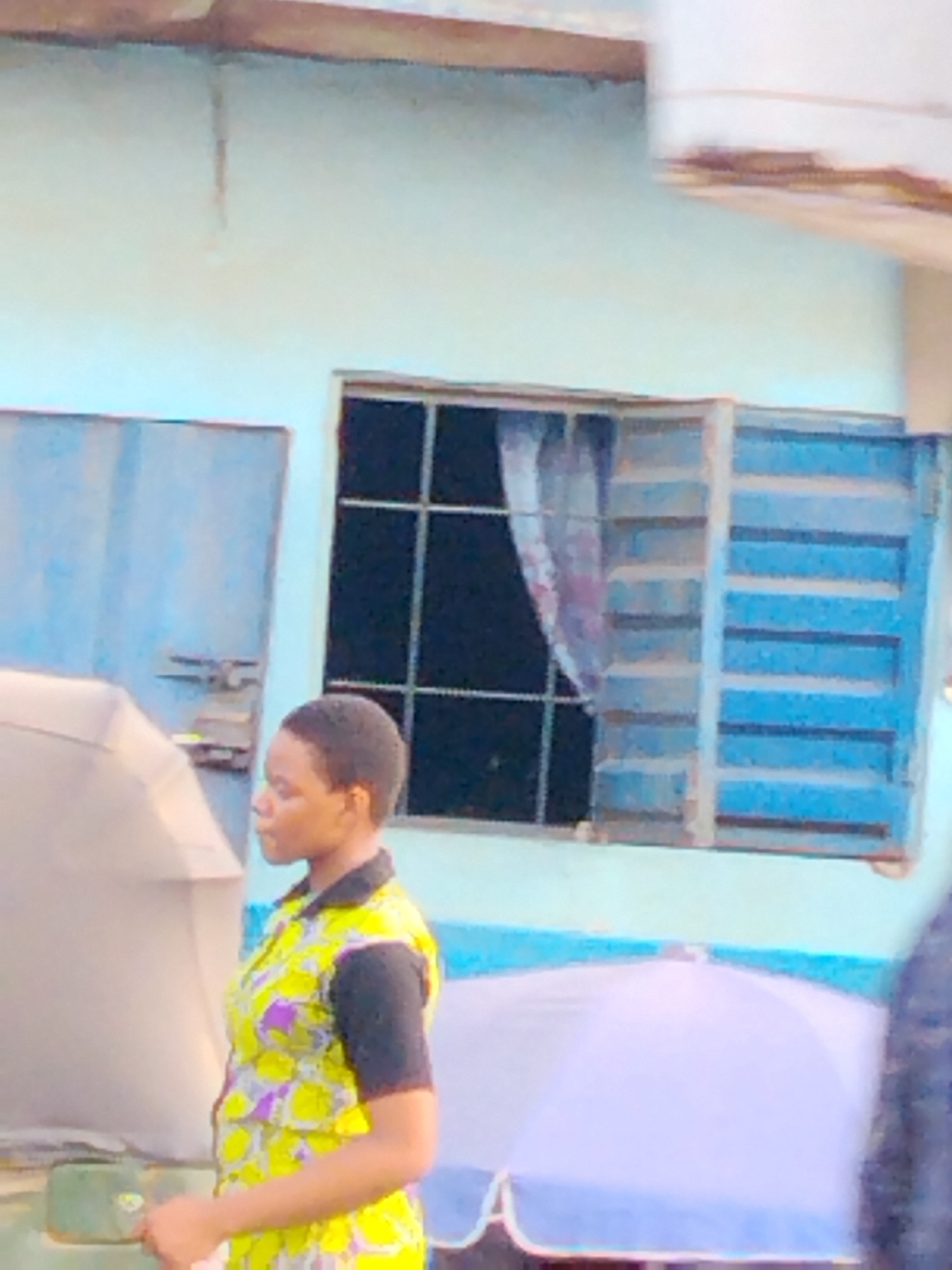 The image shows a woman in a yellow and black patterned dress in front of a light blue building with a barred window and a blue metal door. A partially obscured white and blue umbrella is also visible. The location is likely Aba, Nigeria, based on the dress style and the architecture of the building. There are no other discernible people, interactions, or specific actions occurring.