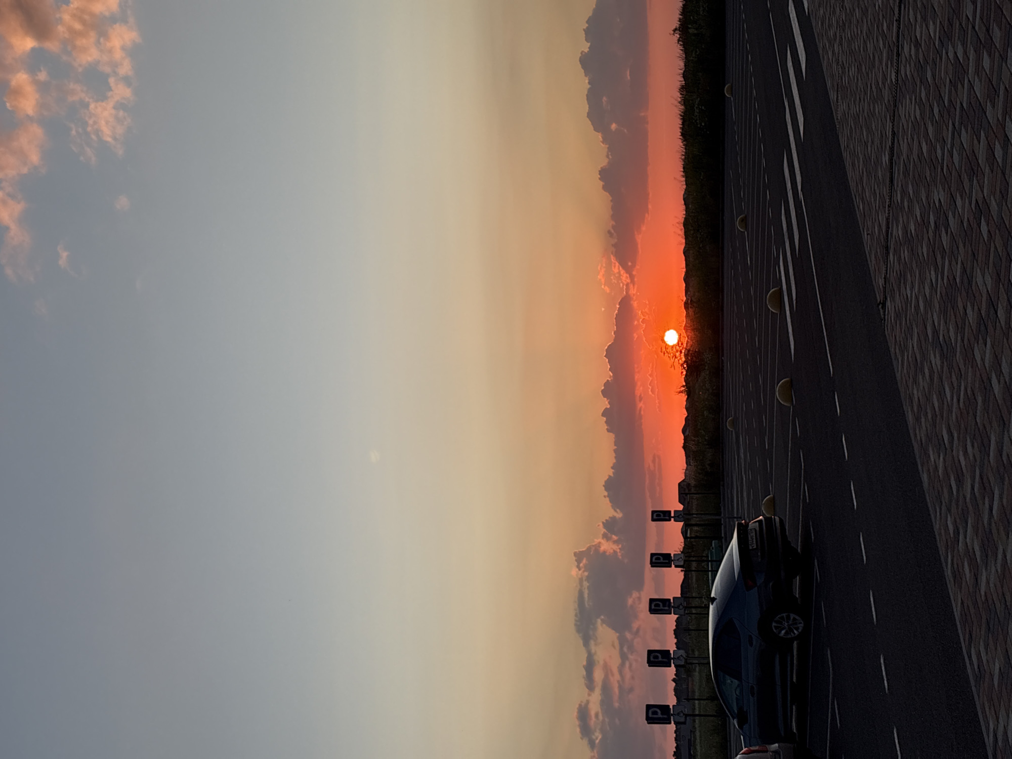 An outdoor, vertical, eye-level shot captures a sunset over a parking lot and a grassy field in Mikhaylovsk, Russia. The sky transitions from a light blue at the top to a pale orange and then a vibrant red near the horizon. Fluffy clouds are tinged with pink and orange by the setting sun. The sun itself, a bright orange orb, is positioned low on the horizon, casting a warm glow.
In the foreground, a dark-colored sedan is parked in a designated spot on an asphalt surface marked with white lines. The parking lot is delineated by yellow bollards. Further back, a row of parking signs with the letter "P" is visible, along with a fence. The grassy field behind the parking lot is silhouetted against the sunset, with some indistinct buildings and trees in the distance. The ground in the immediate foreground is paved with brickwork. There are no people visible in the image.