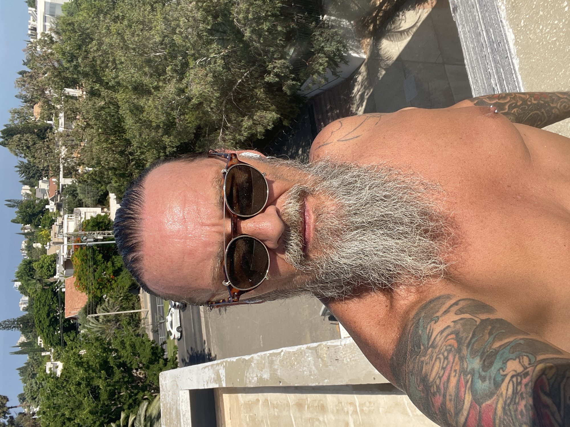 A shirtless man with a long, grey beard and tattoos on his arms is taking a selfie outdoors, likely on a rooftop in Tel Aviv, Israel. He is wearing round, tortoiseshell sunglasses and white earbuds. The background shows a view of urban buildings, trees, and a street with a white car driving by. The scene suggests a sunny day with a clear blue sky.