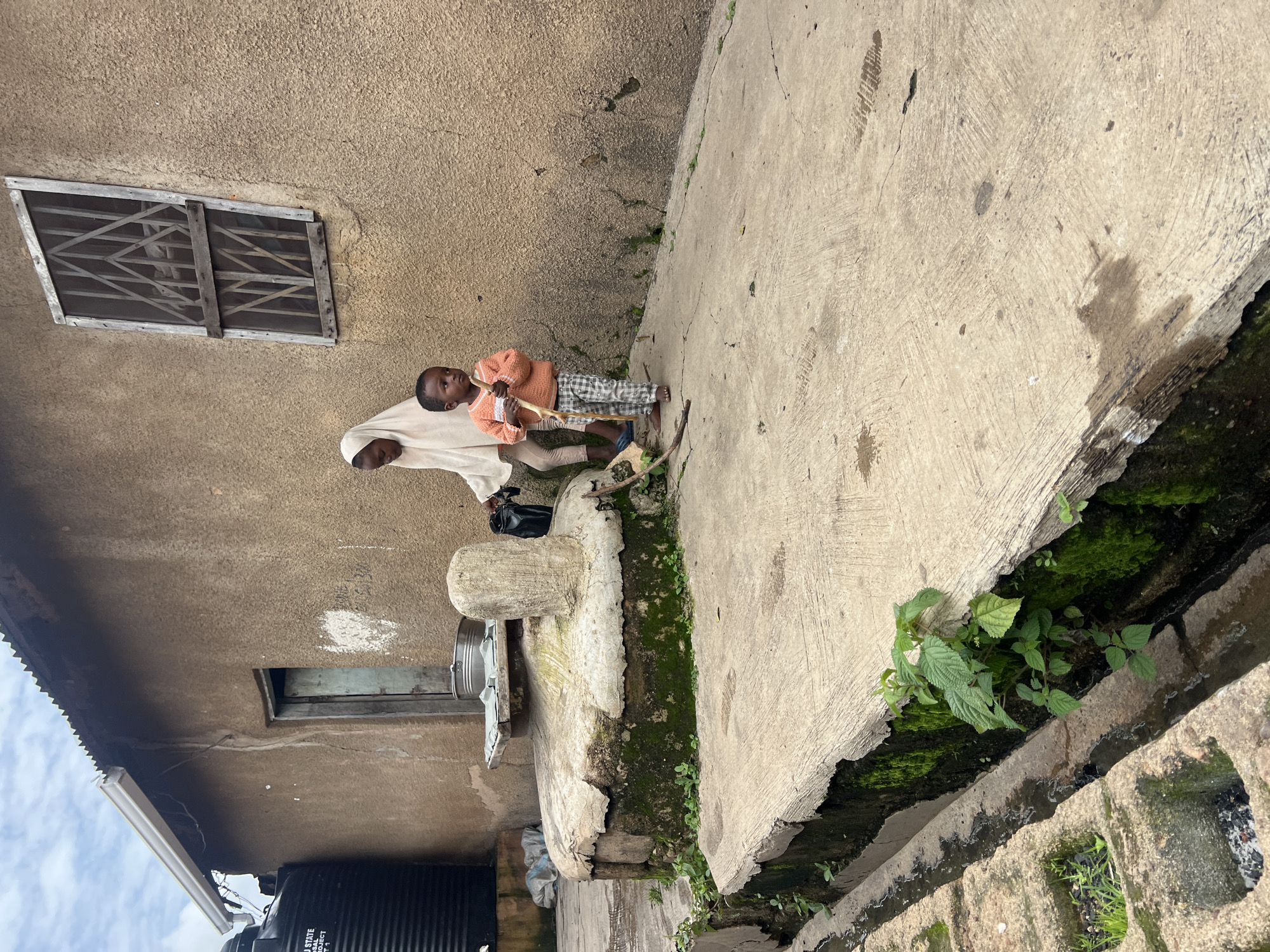 In Jos, Nigeria, two children stand on a concrete pathway next to a weathered, tan-colored building. A young boy, wearing a checkered pattern on his pants and an orange sweater, holds a stick. Beside him, an older child, covered by a light-colored hijab, holds a black bag. The building features a barred window and a metal water tank on the left. A circular, moss-covered structure with a stone pillar stands near the children. A drainage ditch runs along the edge of the concrete path, with visible moss and greenery.