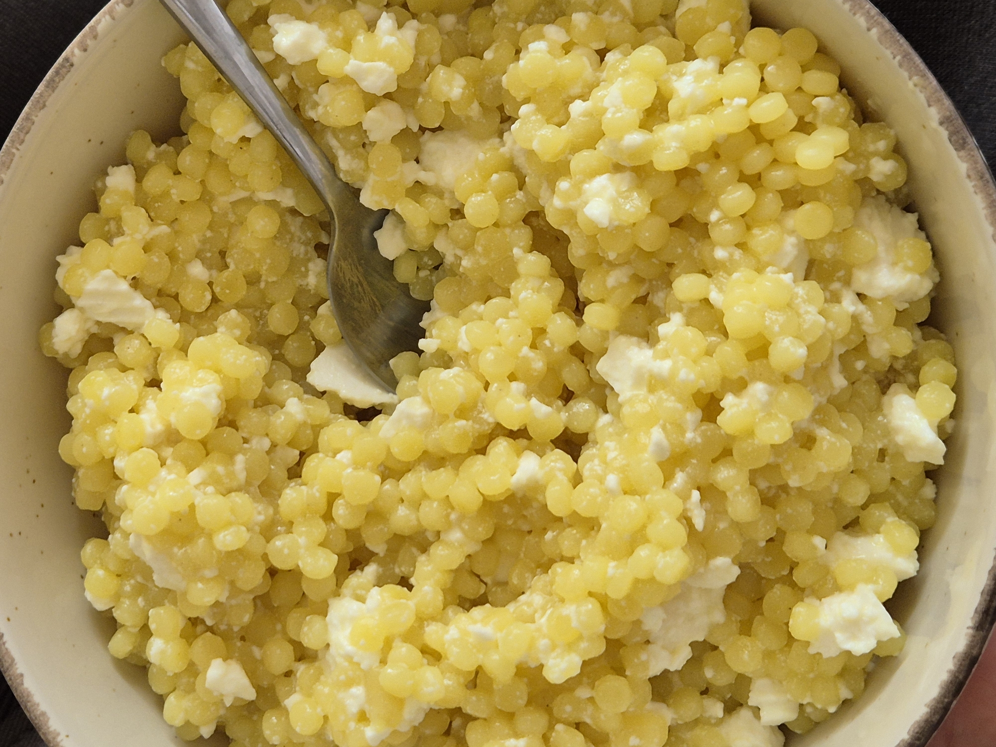 A close-up overhead shot shows a bowl of food, specifically couscous pasta mixed with what appears to be soft white cheese, likely ricotta or cottage cheese. A silver spoon rests in the center of the bowl. The bowl is off-white with a slightly textured rim. The food is photographed under natural lighting.