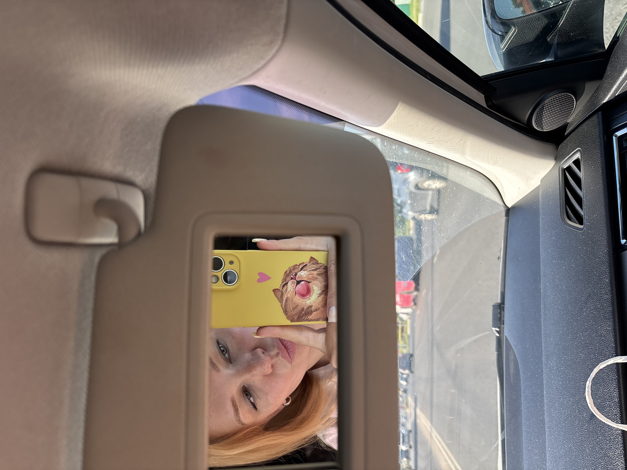 A woman with blonde hair takes a selfie in a car's visor mirror. She is wearing a pink shirt and has a playful expression, puckering her lips. She is holding a yellow smartphone with a cat-themed case. The car's interior, including the dashboard and part of the passenger side, is visible. Outside the car, a road with other vehicles and some buildings can be seen. The location is the city of Zhukovo, Russia.