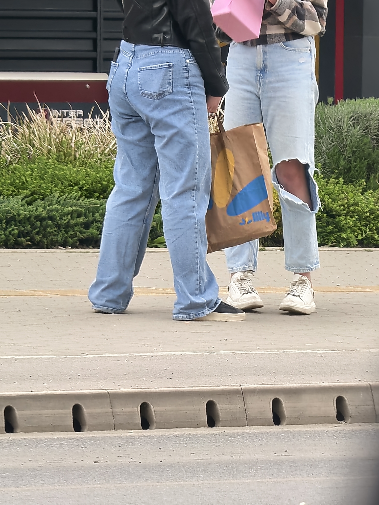 Two individuals are pictured from the waist down, standing on a concrete sidewalk next to a street curb. The person on the left wears a black jacket over light blue, straight-leg jeans and black slip-on shoes. The person on the right is dressed in a plaid shirt, light blue distressed jeans with a large tear on the right knee, and white sneakers.
The person on the right holds a brown paper shopping bag with a blue and yellow logo, and a pink box in their hands. The logo on the bag appears to be "3.J.My". The scene suggests a daytime setting, with natural light illuminating the subjects and their surroundings.
In the background, lush green bushes and tall grass are visible, with parts of a building behind them. A red and white sign on the building has partially visible text, which appears to read "INTER" and "TREN", possibly part of a business name. The concrete curb in the foreground features a series of three oval-shaped drainage holes, indicating a street or parking area adjacent to the sidewalk. The activity suggests the individuals are either walking or paused during a shopping trip.