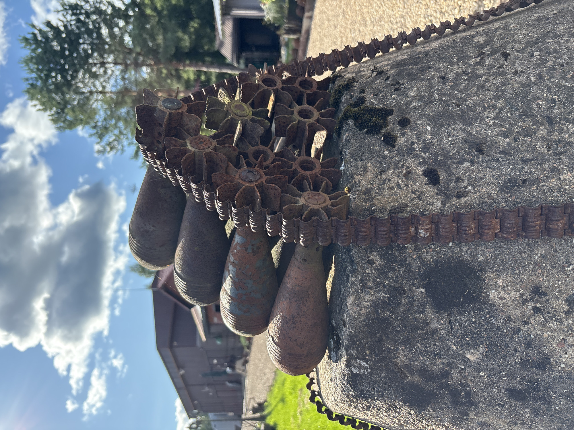 In Zhalybnya, Russia, on a sunny day with scattered clouds, a pile of old, rusty artillery shells rests on a large concrete base. The shells, with their ribbed surfaces, are arranged in a stack, with a long, rusted metal chain draped over them. The chain appears to be a tank track. The concrete base is weathered and mossy in places, suggesting it is outdoors. In the background, a building with a dark roof and wooden siding is visible, along with trees and a gravel path. The scene appears to be part of an outdoor display or memorial.