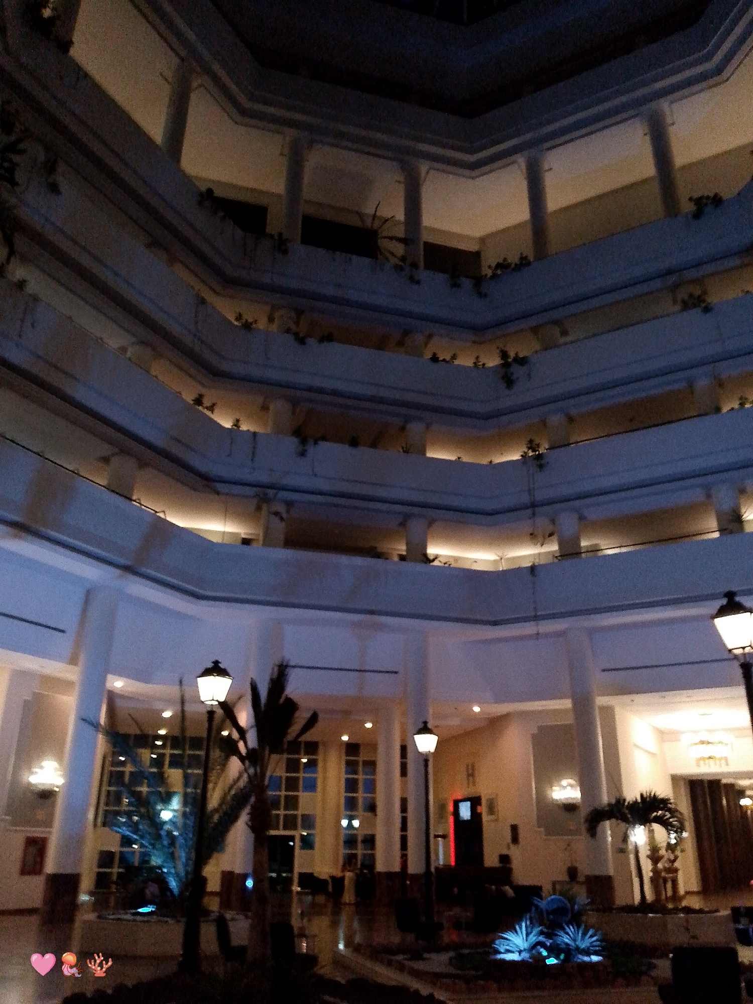 The image depicts the interior of a multi-story building, likely a hotel, located in Pedro González, Venezuela. The structure features a central atrium with multiple levels visible, each with balustrades and columns. Plants are visible along the balustrades. The ground floor contains a lobby with seating areas and decorative plants, illuminated by streetlights and other light fixtures. Tall windows are also visible. People are present in the seating areas.