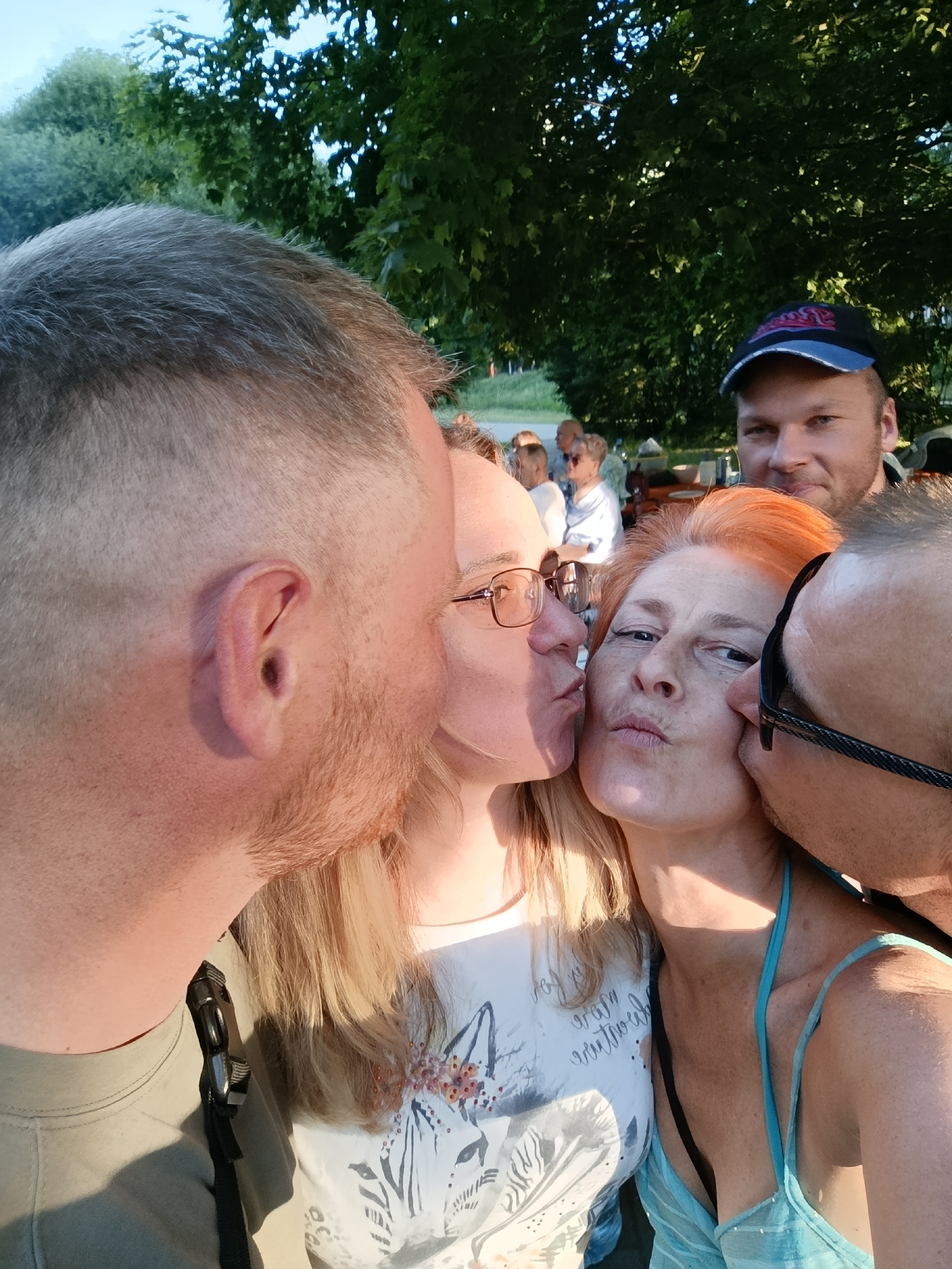 The image depicts a close-up, candid outdoor scene featuring five individuals, with three central figures engaged in an intimate interaction. The setting is an unspecified outdoor location within Moscow, Russia, characterized by extensive green foliage from trees and bushes under what appears to be late afternoon or early evening natural light.
In the foreground, a light-skinned male with short, graying hair and an ear visible on the left is positioned with his face close to a light-skinned blonde female, suggesting an act of cheek-kissing. This blonde female wears glasses and a white t-shirt with a dark patterned design. She is also positioned with her face close to a central light-skinned female with bright orange-red hair, indicating a second act of cheek-kissing. The central red-haired female wears a light blue halter-neck top, has her eyes slightly open, and her lips pursed in a pout. On the right, a third light-skinned male, partially visible with dark hair, light stubble, and glasses, is positioned with his face close to the central red-haired female, completing a third act of cheek-kissing.
In the mid-ground, behind the central red-haired female, a fourth light-skinned male wearing a blue and black baseball cap with an indiscernible logo is visible. He has a beard and is looking towards the camera with a slight smile. Further in the blurred background, several other light-skinned individuals are discernible, seated or standing around what appear to be picnic tables or similar outdoor furniture, indicating a social gathering. The distant background shows more green vegetation and a light sky.