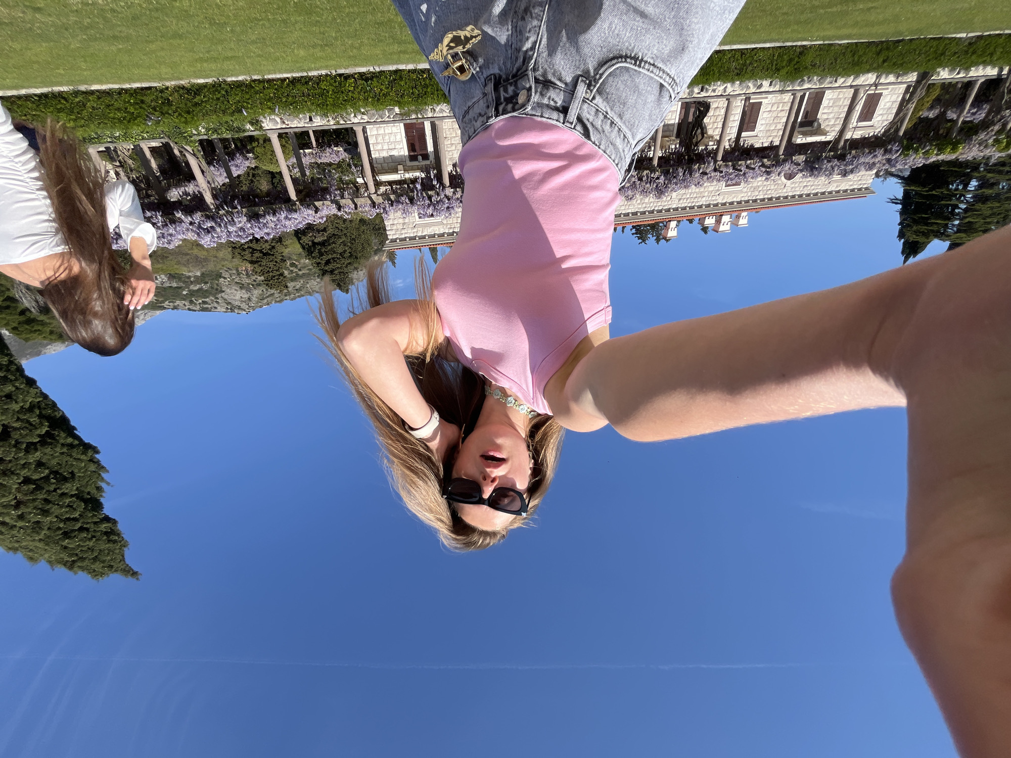 The image shows two women at a park in Sveti Stefan, Montenegro. One woman is wearing a pink tank top, jeans, and sunglasses. She is taking a selfie. The other woman is wearing a white top and is visible in the background. A building with purple flowers growing nearby is visible in the background. The sky is blue and there is a tree visible on the left side of the image.