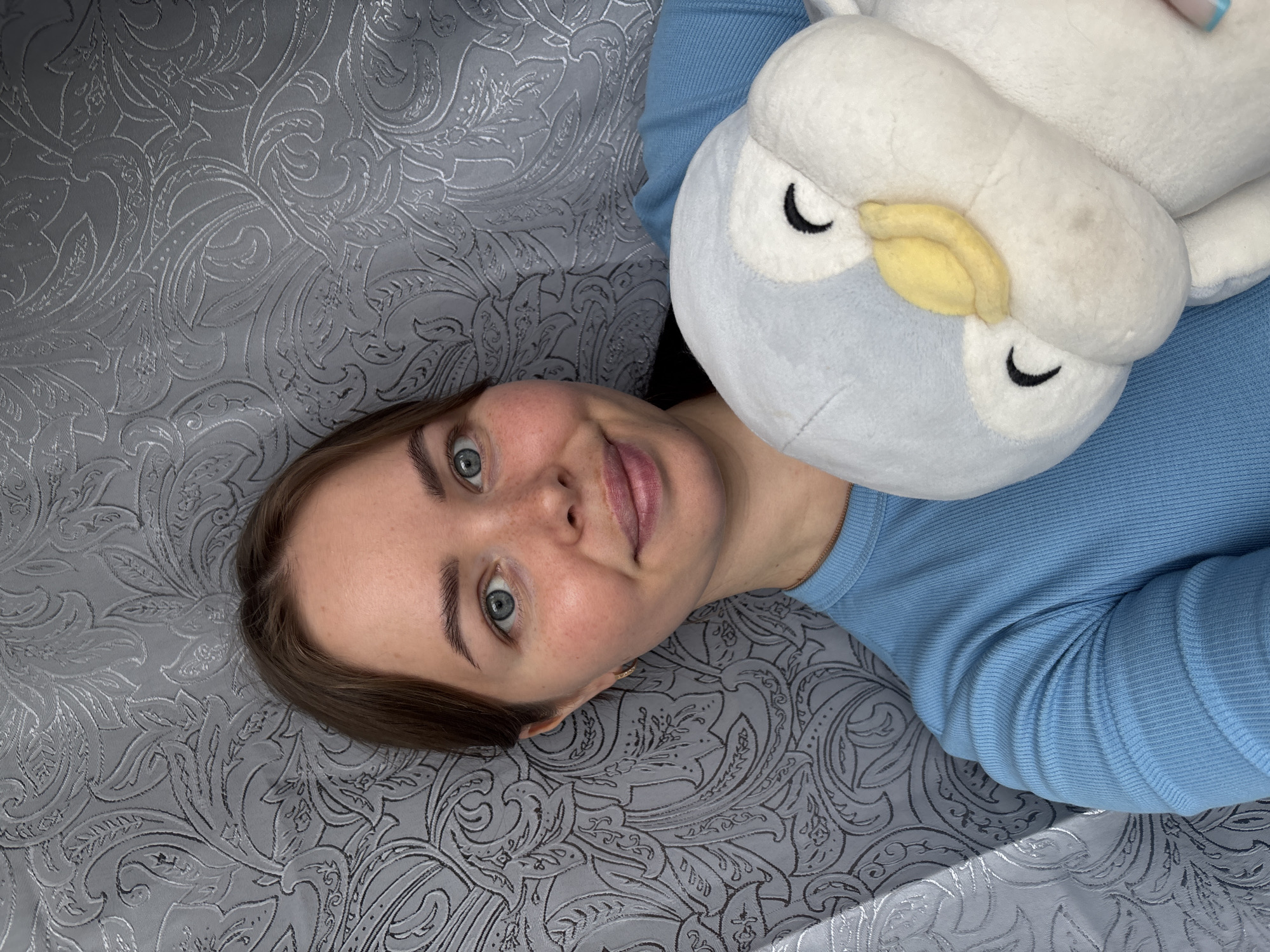 A young woman is depicted in a close-up, high-angle shot, lying on her back and looking directly at the camera. She has light brown hair, blue eyes, and a neutral expression with a faint smile. She is wearing a light blue, ribbed, long-sleeved top. To her right, snuggled close to her head, is a soft, light blue and white plush toy, resembling an owl or a bird, featuring black crescent-shaped closed eyes and a small yellow beak. Both the woman and the plush toy are resting on a textured gray or silver surface, likely a bedspread or blanket, which is adorned with an intricate, embossed floral or paisley pattern that exhibits a subtle sheen. The lighting is soft, suggesting an indoor setting. There is no visible text in the image.