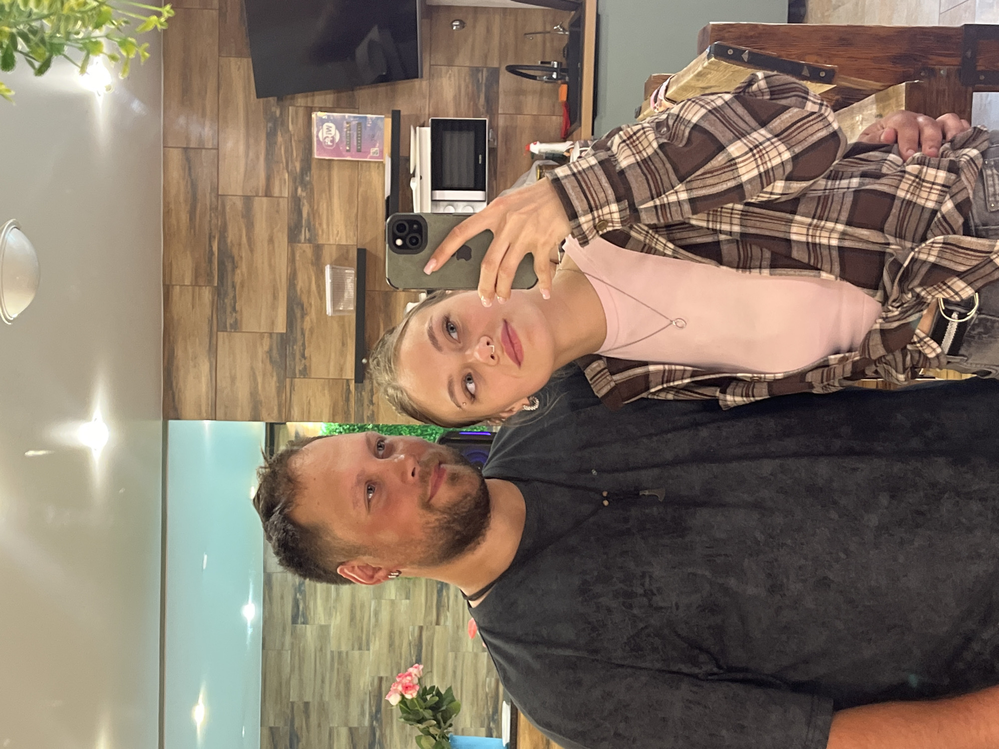 The image captures a selfie taken by a fair-skinned young woman with blonde hair, wearing a pink top and a brown plaid shirt, holding a smartphone with an olive green case. She has a nose piercing and a necklace. Behind her, a fair-skinned man with a beard is visible, wearing a dark grey t-shirt. The backdrop appears to be a room with wooden-paneled walls, a television, and a microwave. There is also a decorative plant in the foreground. Based on additional information, the location is Chaikovsky, Russia.