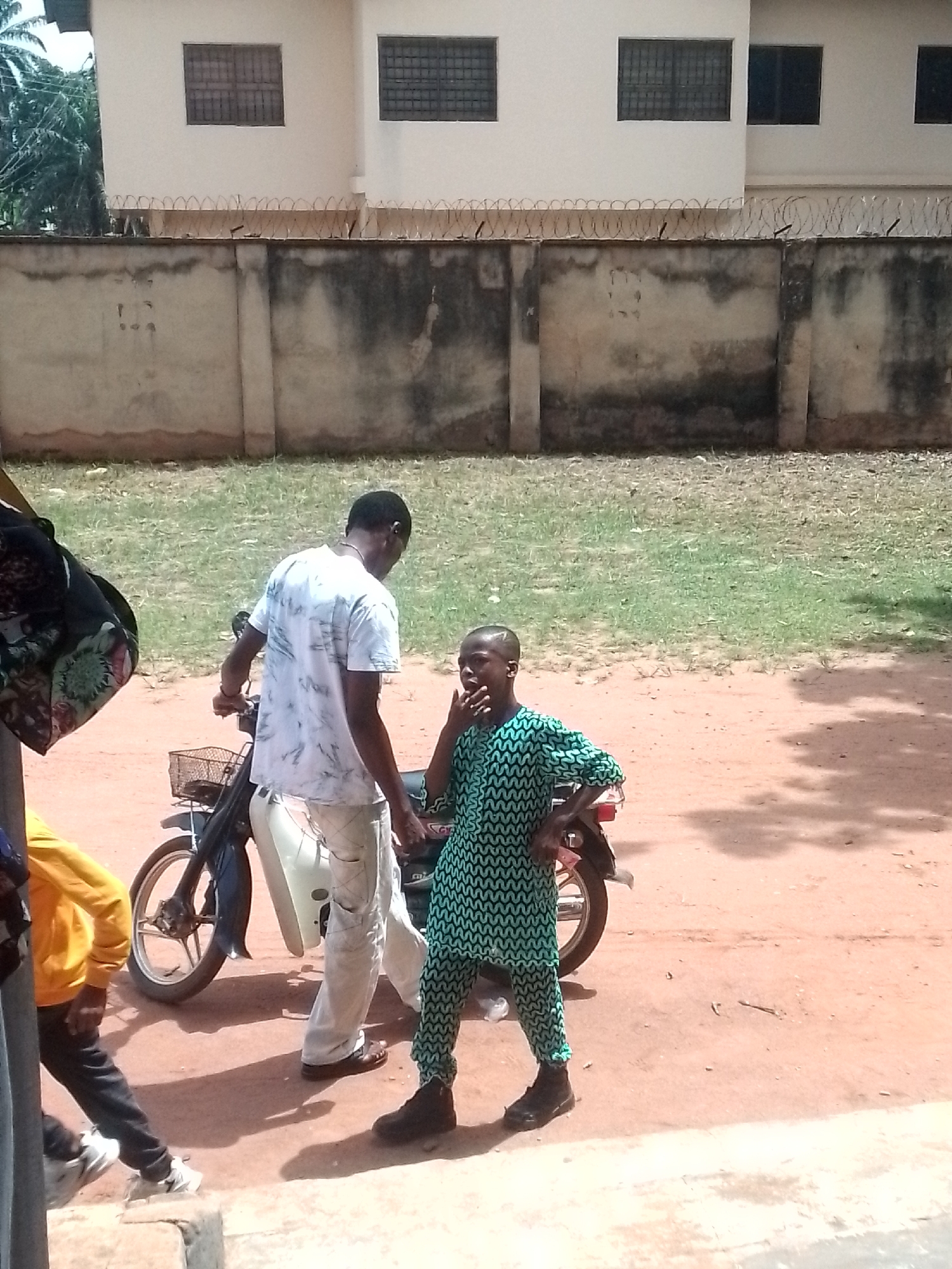 The image shows two people next to a motorbike in Orlu, Nigeria. One person is standing beside the bike, wearing a patterned green outfit, with their hand to their mouth. The other person is standing behind the bike, wearing a white shirt and light-colored pants. A person wearing a yellow jacket is seen partly in the bottom left of the image. The motorbike has a basket on the front. The background includes a building with barred windows and a barbed wire fence. A grassy area is visible between the fence and the building. The ground surface appears to be dirt. The scene is outdoors in daylight.