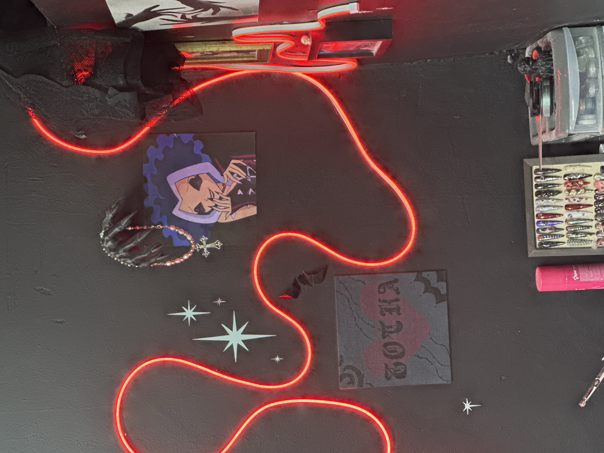 An indoor, overhead shot captures a dark, textured wall in what appears to be a personal space or a creative studio in Novosibirsk, Russia. The scene is dominated by a vibrant, snaking red neon light strip that crisscrosses the wall, providing the primary illumination.
Various decorative items and tools are affixed to or placed against the wall. Towards the top left, black, netted fabric is draped near the neon light. Below it, a square artwork depicts a character with purple, wavy hair and heart-shaped eyes, striking a pose with stylized hands. Nearby, a beaded bracelet with a silver cross charm hangs. Further down, several white star decals of varying sizes adorn the wall. Another square canvas, dark gray with red accents, features the word "ТОЧКА" (TOCHKA) in a Gothic-style font, possibly accompanied by a cherry or heart design. A small, black bat cutout is positioned above this canvas, near the neon light.
On the upper right, an electronic device, possibly a timer or counter, is mounted to the wall, partially obscured. Below it, a framed display case showcases numerous colorful, elongated objects, resembling nail art tips or fishing lures. A pink spray bottle and a silver, glittery pen or tool are also visible on the right side of the frame.
The overall aesthetic is dark and eclectic, combining elements of alternative or gothic styles with crafting or beauty accessories. The artificial red neon light creates a moody and dynamic atmosphere, suggesting the image was taken indoors, possibly at night or in a dimly lit environment.