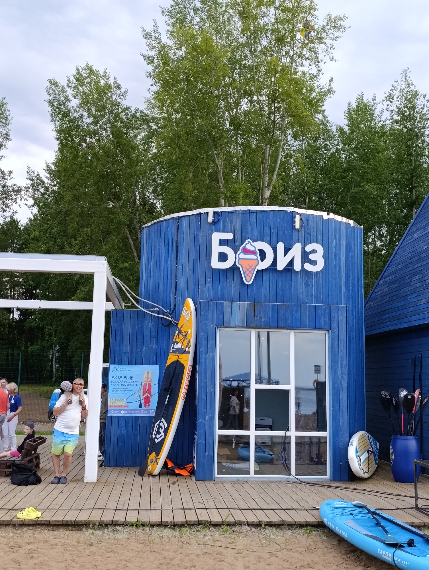 This is a medium shot, eye-level photograph taken outdoors on a sandy beach, likely in Chaikovsky, Russia, under an overcast sky. The scene features a bright blue wooden building with a prominent sign displaying the Cyrillic word "Бриз" (Breeze) and a stylized ice cream cone. In front of the building, a wooden deck is laid out.
Several people are present. On the left, a man in a white t-shirt and colorful shorts carries a child on his shoulders, both smiling. Other individuals are visible in the background near the building and further back.
Various objects related to water sports are scattered around. A yellow and orange stand-up paddleboard (SUP) leans against the blue building, its brand name "GO BOARD" visible. Another blue SUP lies on the sand in the foreground, with oars resting on it. A blue barrel filled with SUP oars stands near the building, and a white inflatable disc-like object is propped up against the wall. An orange life vest is also visible on the deck.
A poster is attached to the blue building with Cyrillic text that includes "АКВА-РЕЙВ" (Aqua-Rave) and instructions for a "stand up on the dock, change reality" event. The overall atmosphere appears relaxed and summery, with people enjoying a day at the beach or a waterfront activity. The time of day is difficult to ascertain precisely due to the overcast weather, but it appears to be daylight hours.