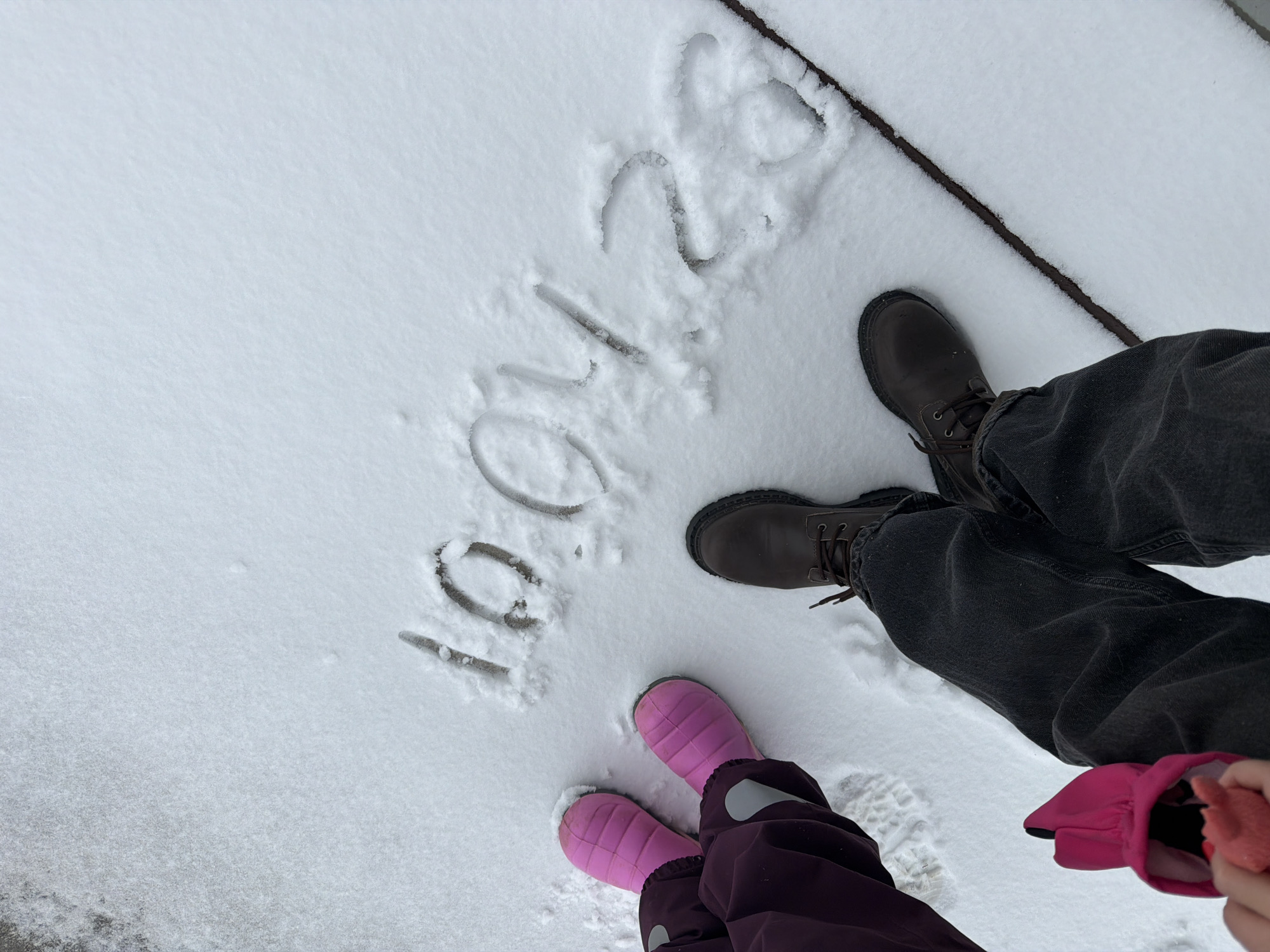 The image shows a snowy outdoor scene. The numbers "10.01.24" are written in the snow. Two pairs of feet are visible. One pair wears brown boots and dark jeans. The other wears pink snow boots and purple snow pants. A pink glove is also visible. The location cannot be determined from the image.