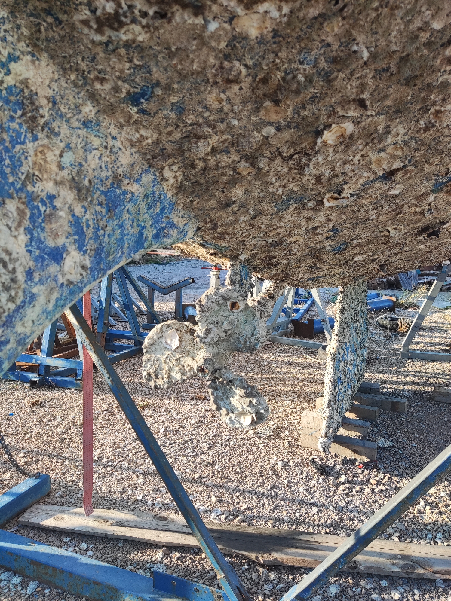 The image shows the underside of a boat, encrusted with marine growth and supported by blue metal stands on a gravel surface. A propeller, also heavily fouled with growth, is visible. In the background, other boats on stands and various nautical equipment can be seen. The scene appears to be a boatyard in Bar, Montenegro, likely during maintenance or storage. The lighting suggests it is daytime, with sunlight casting shadows.