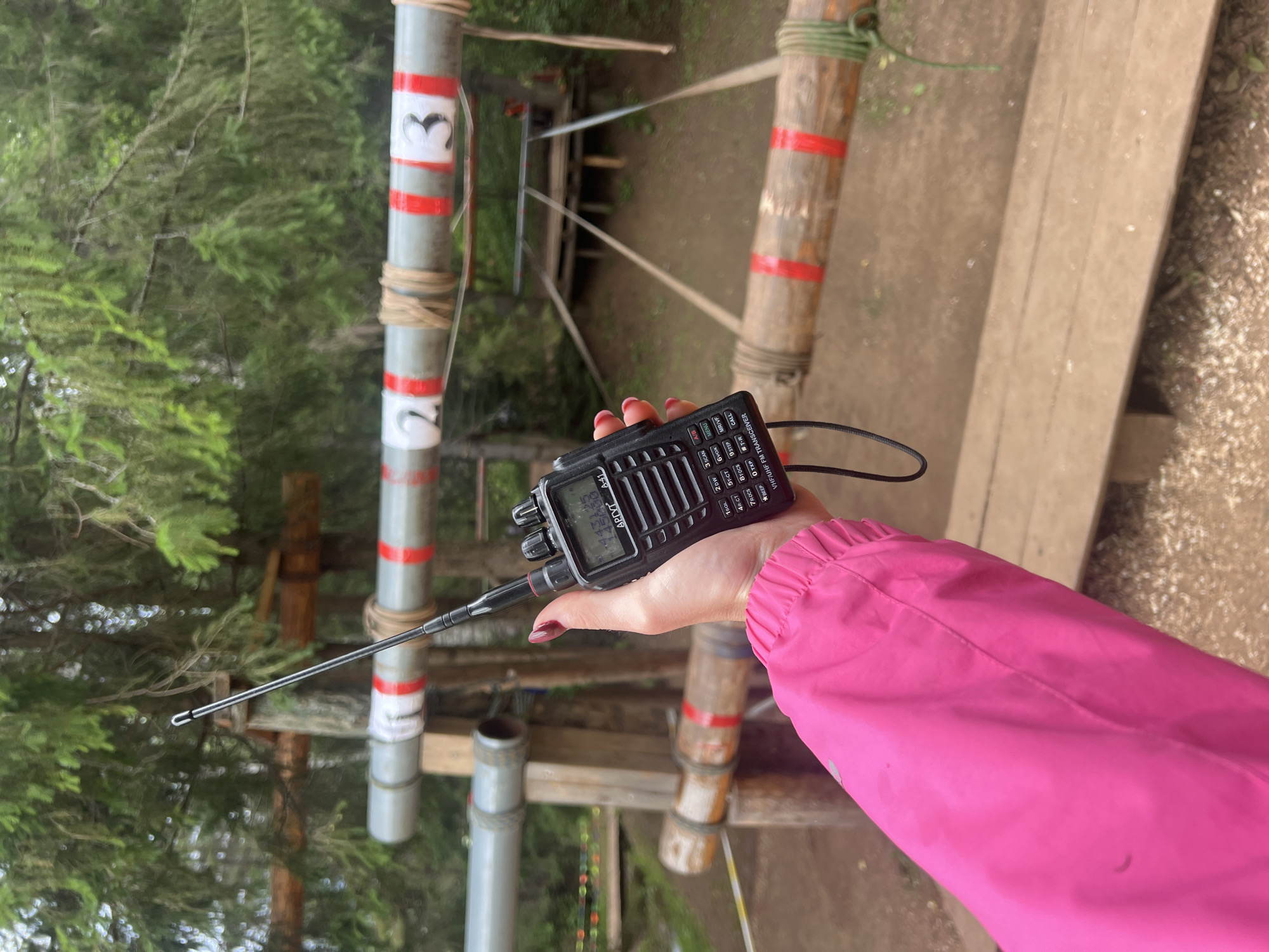 The media file shows a close-up of a person's hand holding a black walkie-talkie. The walkie-talkie is a VHF/UHF FM transceiver with a small display screen and several buttons. The screen displays "A-41" and some numbers and letters that are difficult to read. The buttons are labeled with functions like "SCAN," "MENU," "CALL," and "BEEP." The person's hand has dark red nail polish. They are wearing a bright pink jacket, the sleeve of which is visible on the right side of the frame.
The background suggests an outdoor setting, possibly a forest or park, with trees and wooden structures. There are vertical poles in the background, one of which is grey with red and white bands and a symbol that resembles a stylized heart or a number "3." Another pole is made of bamboo with red bands. The ground appears to be made of dirt and gravel. The lighting suggests it is daytime, and the overall atmosphere is casual, perhaps indicating an outdoor activity like hiking, a game, or work. The presence of the walkie-talkie implies communication is taking place or is about to take place. The location context provided is Сергино, Russia.