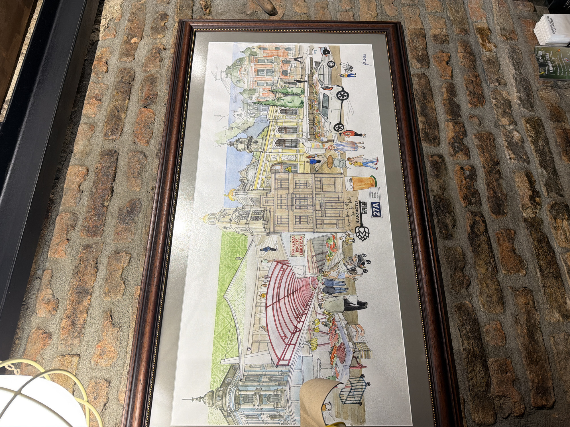 This media file showcases a framed, panoramic watercolor illustration depicting a vibrant street scene, set against a rustic brick wall. The illustration captures a lively urban environment, likely in Varna, Bulgaria, given the context.
The scene is dominated by ornate, multi-story buildings with architectural details, including domes and decorative facades. On the left, a grand building features a prominent entrance with a red arched canopy, beneath which people are gathered. Further along the street, another building with a green dome and intricate details is visible.
Numerous stylized people populate the street and sidewalks. They are engaged in various daily activities: walking, conversing, pushing strollers, and shopping at outdoor market stalls filled with colorful fruits and vegetables. Two white cars are parked along the street, suggesting a busy urban thoroughfare. Outdoor tables and chairs are also visible, indicating cafes or restaurants.
Notable details include the bright, daytime illumination, and the distinct architectural style. Several signs with Bulgarian text are visible: one red sign under the canopy reads "ПРИ НУЖДА ТЪРСЕТЕ СПАСИТЕЛИ" (In case of need, seek rescuers/lifeguards). A vertical sign advertises "Ключето БИРА БАР" (Klyucheto Beer Bar) with the number "27A" and a hop logo. Additional text, "КЛЮЧОВА СЦЕНА всички клиенти са ключови" (Key Scene, all clients are key), and a signature "R.2025" (possibly Y.P. 2023 based on closer inspection, or related to the bar's establishment/anniversary) are also present. The overall artwork style is a charming, somewhat whimsical rendering of a bustling city center.