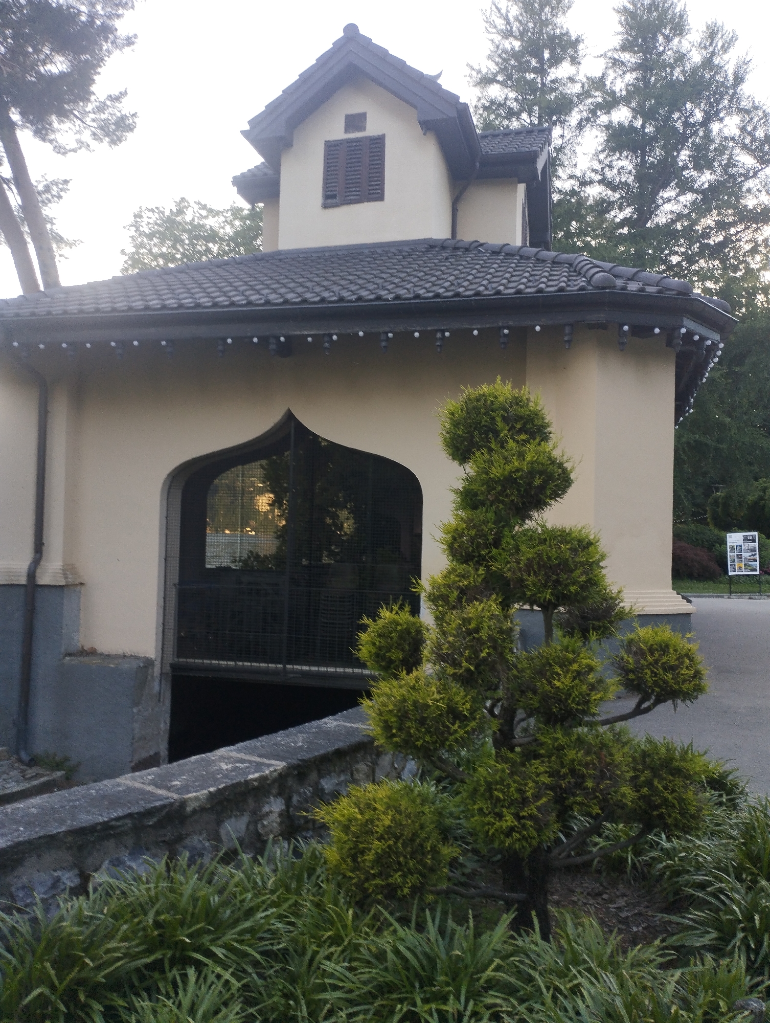 The image depicts the exterior of a building with a distinctive arched entrance, possibly a gatehouse or entrance to a park, set against a backdrop of trees and an overcast sky. The building features a light-colored facade with a dark, tiled roof. A window with dark shutters is visible on the upper story. In the foreground, a neatly trimmed, conical evergreen tree stands to the right of the entrance, with a low stone wall and lush green foliage in front of it. To the right of the building, a sign with multiple images and text is visible, suggesting it might be an information board. The overall setting appears to be an outdoor public space, likely a park or garden within the city of Lugano, Switzerland, given the characteristic architecture and landscaping.