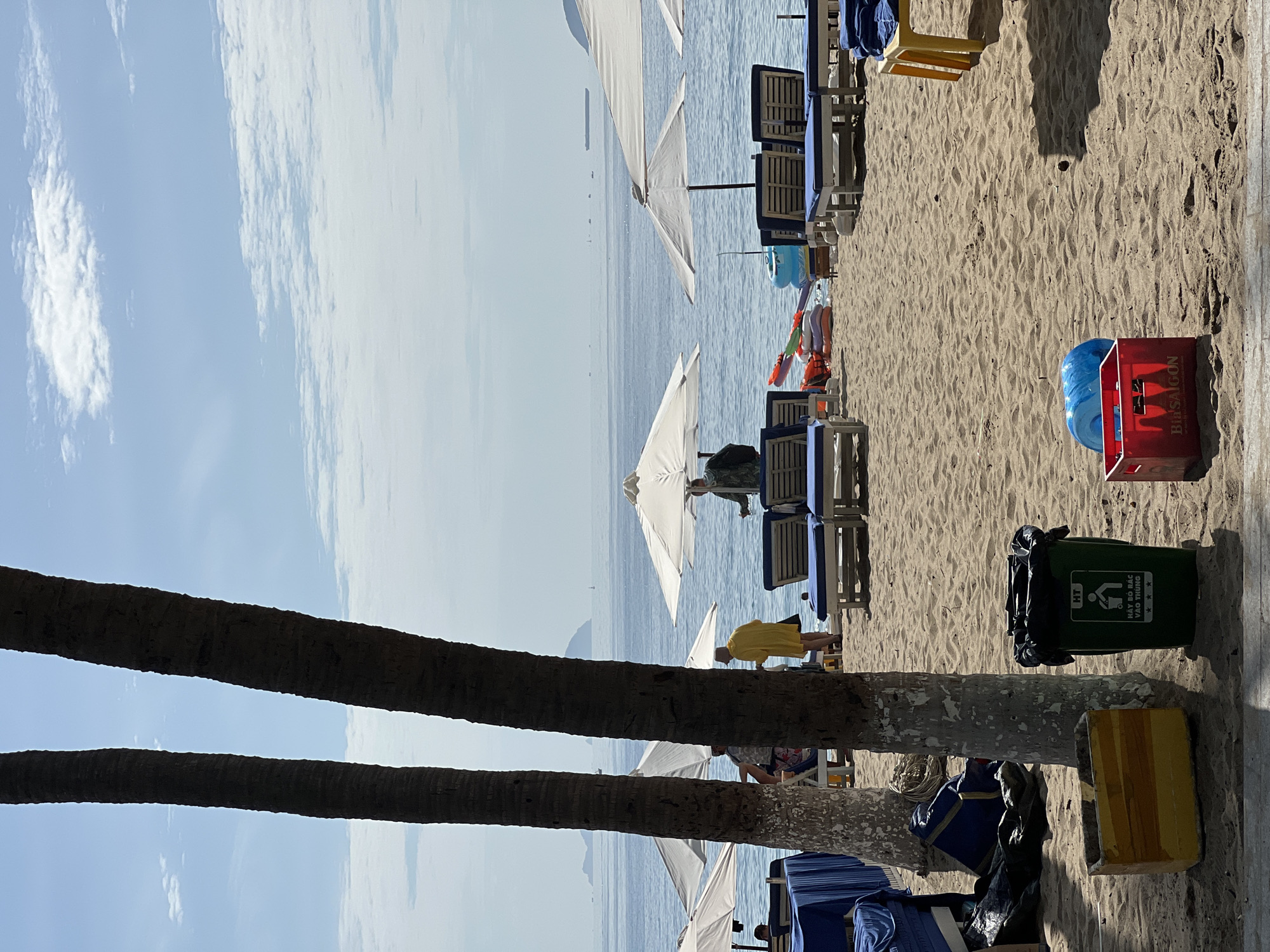This is a photograph taken on a sunny day at a beach in Nha Trang, Vietnam. The foreground shows a sandy beach with two palm trees partially obscuring the view. To the left, there are blue cloths spread over what appear to be tables or loungers, and a blue bag is on the sand. Next to the palm trees, there is a yellow rectangular container and a green trash can with a symbol indicating "HT" and "HAY BO RAC VAO THUNG" (Please put trash in the bin) in Vietnamese. To the right, closer to the water, a red crate with "BIA SAIGON" written on it holds a blue inflatable object.
In the middle ground, several white umbrellas are open, providing shade for blue loungers arranged on the sand. Some people are visible near the loungers. One person in a yellow garment stands near a lounger, facing the water. Another person is seated. Further out, closer to the water's edge, there are some colorful inflatable rings and a blue flotation device.
The background features the calm, blue ocean, with a hazy horizon where several islands or landmasses can be seen in the distance. A few boats and a large cargo ship are also visible on the water. The sky is bright blue with scattered white clouds. The overall scene depicts a typical beach resort atmosphere, likely a public beach or part of a hotel's amenities in Nha Trang.