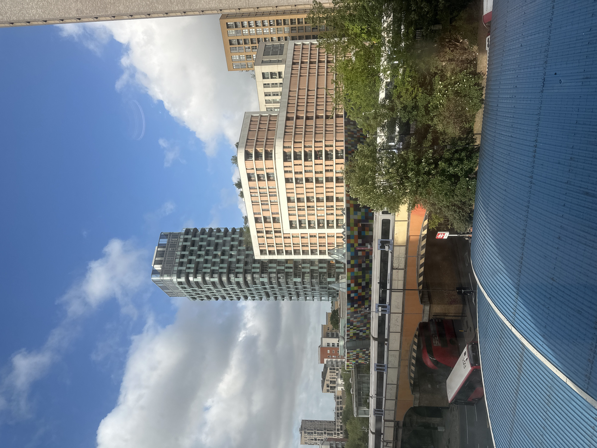 The image shows a view of buildings and transport infrastructure in London, United Kingdom. In the foreground, a corrugated blue structure dominates the right side of the frame. Adjacent to this, a railway bridge in orange tones supports a train. Below, a road tunnel is visible with a red double-decker bus and a white truck. Tall residential buildings stand in the background, with clouds and blue sky partially visible. The image is taken from a slightly elevated position.