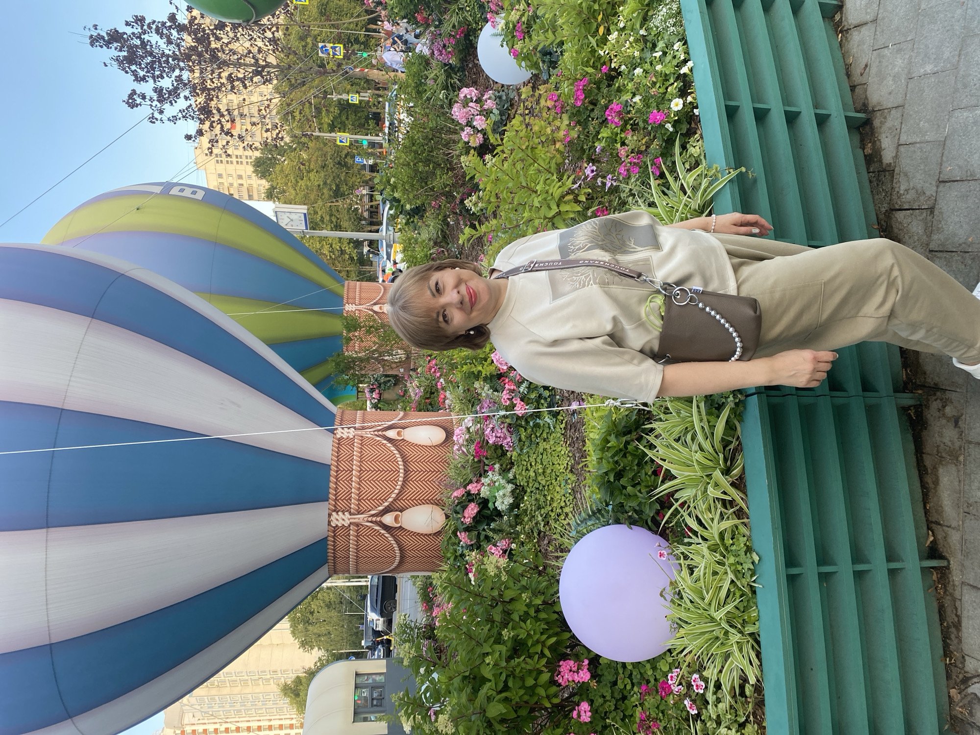 A middle-aged woman with short blonde hair and red lipstick stands in front of a flowerbed. She is wearing a light brown crewneck sweatshirt and matching light brown pants. She has a brown crossbody bag with a silver beaded strap slung over her shoulder. In the background, two large hot air balloons are visible. One is blue and white striped, and the other is green and blue. The flowerbed is filled with pink, white, and green flowers and foliage. There are also two large spherical decorations, one purple and one white, placed within the flowerbed. The scene appears to be in a city park or public square, with buildings and traffic visible in the distance. This location is in Moscow, Russia, specifically on Poklonnaya Hill.
