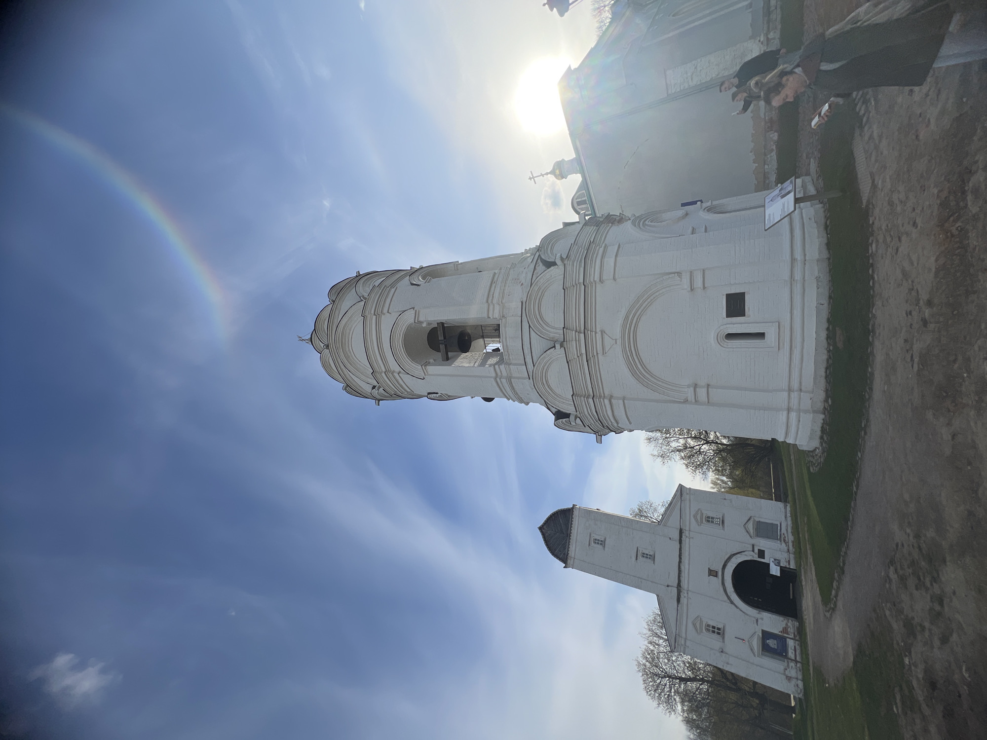 The image depicts a low-angle shot of the Church of the Ascension in Kolomenskoye Park, Moscow, Russia. A rainbow arches across the left of the sky. The white stone bell tower of the church is in the foreground. In the background is the church and ground with people.