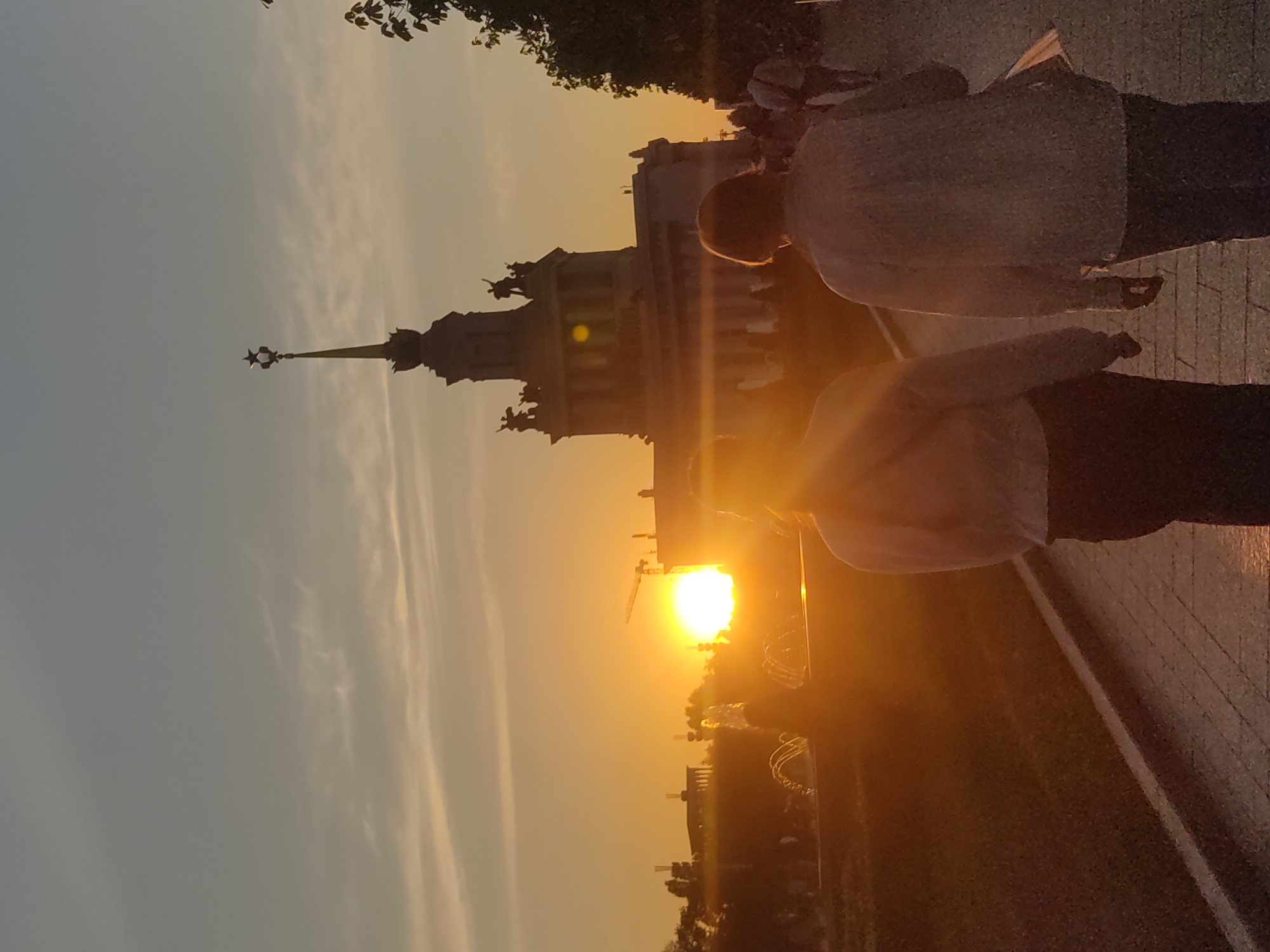 Here's a description of the image:
The image shows a sunset view in Moscow, Russia. A prominent landmark, identifiable as one of the Seven Sisters skyscrapers (likely the Kudrinskaya Square building), is silhouetted against the setting sun. Two people are standing on the right side of the frame, partially obscuring the view. They appear to be observing the sunset. The overall light in the image is warm due to the sunset. A paved walkway is visible in the lower right corner, while trees and other structures are visible in the background near the horizon. The sky transitions from pale blue to orange, reflecting the sunset.