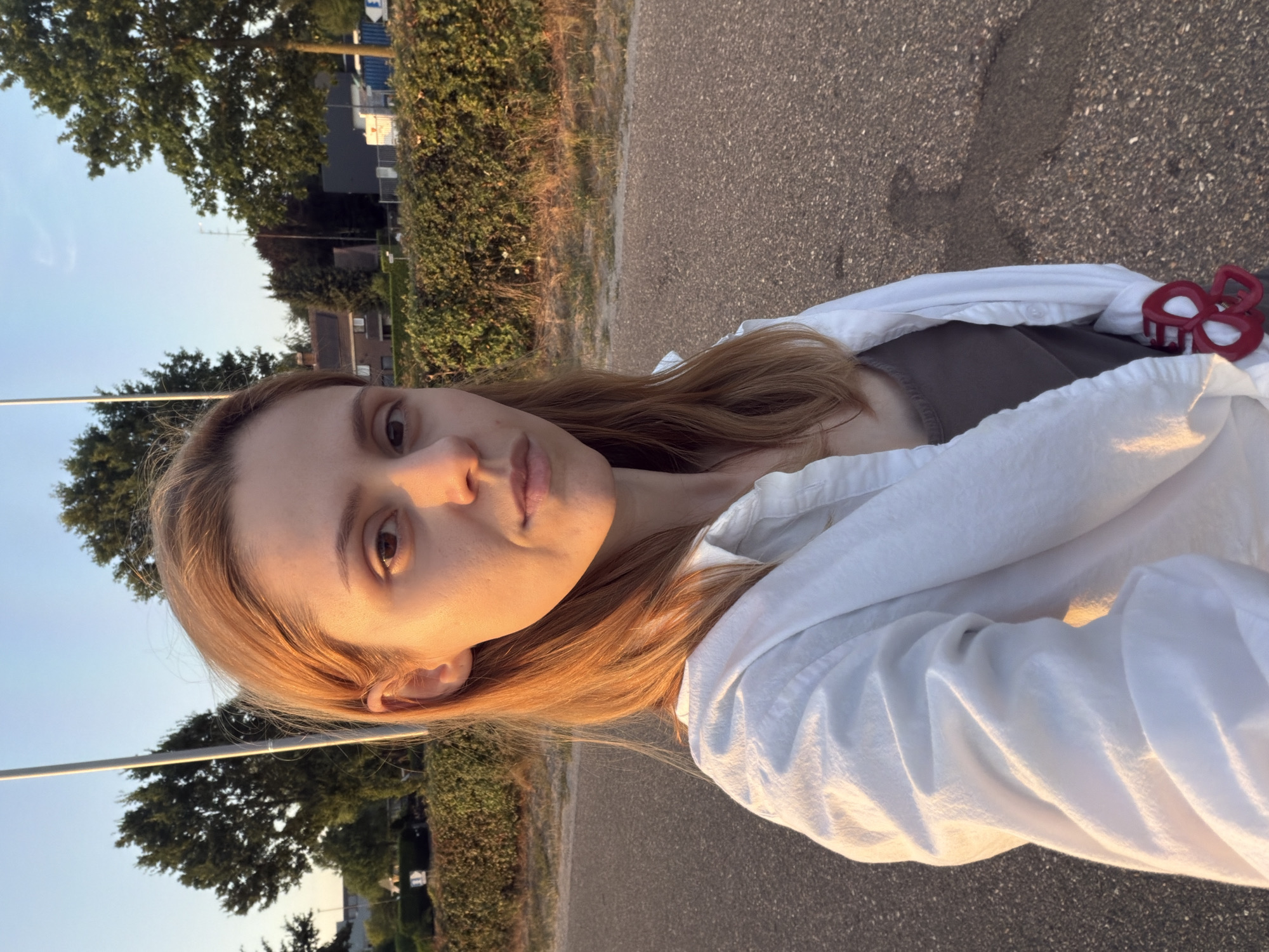 A young woman with auburn hair and fair skin stands outdoors, facing the camera. She is wearing a grey tank top and an open white button-down shirt. Her gaze is directed forward, and her expression is neutral. The background consists of a paved road, a dense green hedge, trees, and some buildings in the distance. The scene is illuminated by the warm light of what appears to be either sunrise or sunset, casting long shadows on the road. The image was captured in Helmond, Netherlands.
