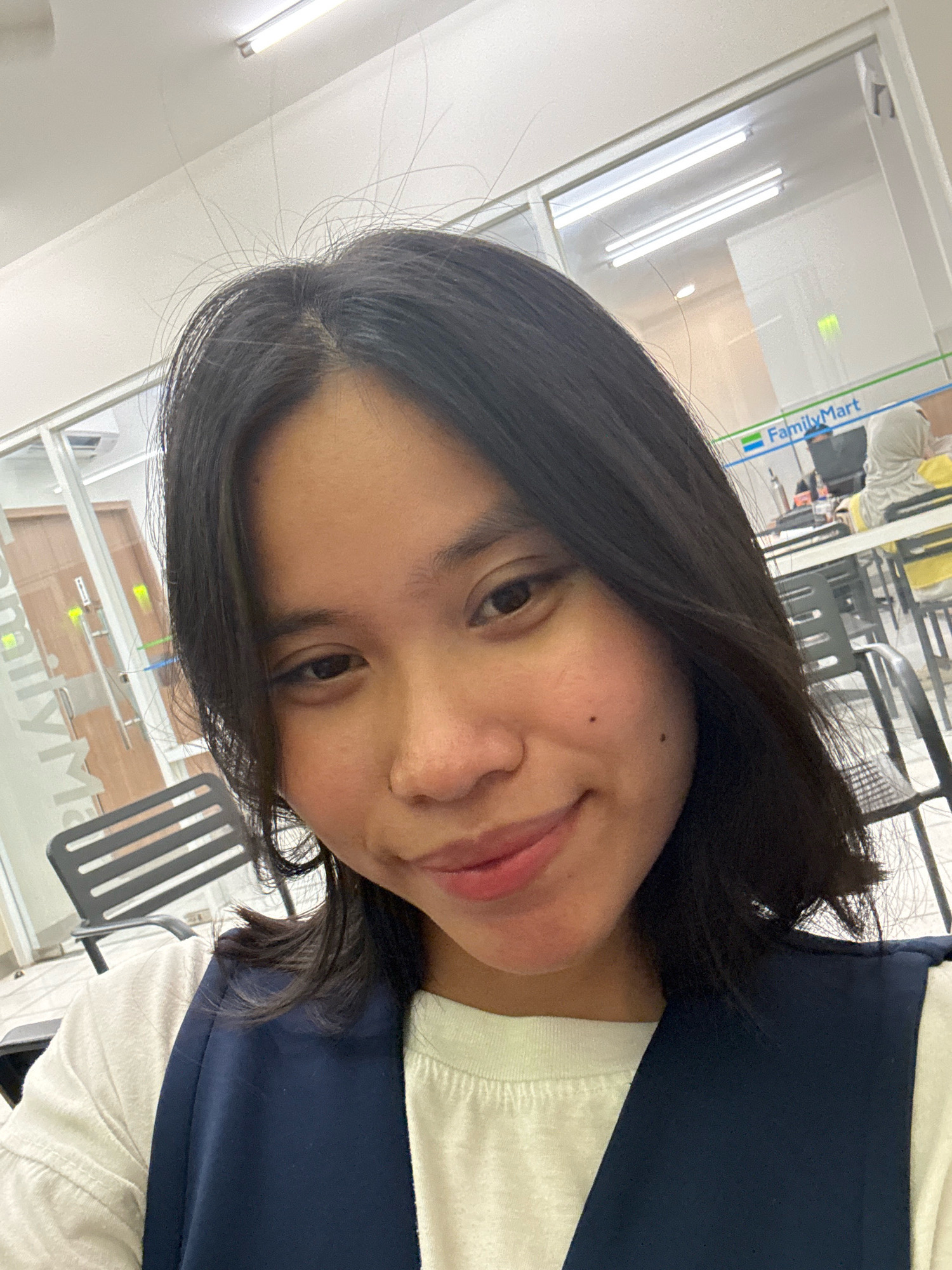 In a selfie taken indoors, a young woman with dark hair and fair skin poses in front of a glass-walled establishment. She wears a white t-shirt and a dark blue vest or apron. Her gaze is directed downwards and slightly to the left, with a subtle smile on her lips. The background reveals a store interior with fluorescent lighting, shelves, and tables with chairs. A sign for "FamilyMart" is visible on the wall, indicating the location is a convenience store, likely in East Jakarta, Indonesia, given the overall context. The setting appears to be a commercial area, with elements of typical store decor and customer activity subtly suggested in the blurred background.