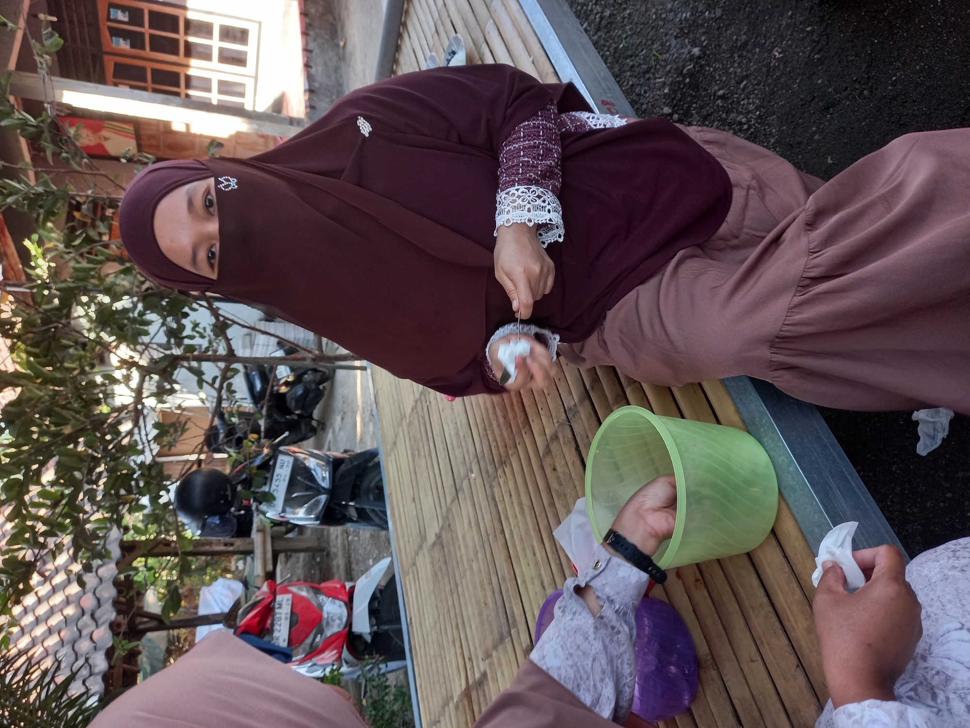 A woman wearing a maroon hijab and niqab sits on a bamboo bench outdoors in Tempos, Indonesia. She holds a small wad of tissue in her left hand. To her left, another person in a pale pink hijab is partially visible, reaching into a purple container. In front of the woman with the niqab is a green plastic bucket, into which her right hand is dipping. Several motorcycles are parked in the background, and a tiled roof structure is visible behind them. The ground is covered with gravel.