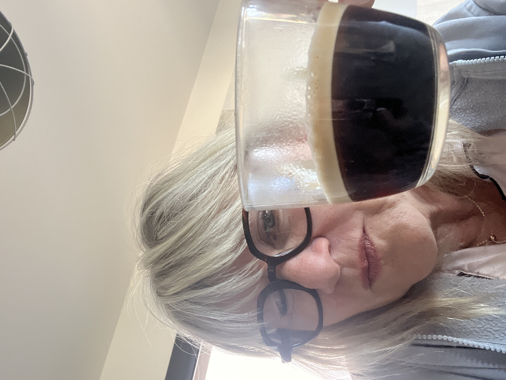 A close-up, overhead shot shows a woman holding a clear glass of coffee with a foamy crema. She has blonde hair and is wearing glasses. She is looking down at the coffee. The background is blurred and appears to be an indoor setting. The specific location in Chambœuf, France, is not discernible from the image.