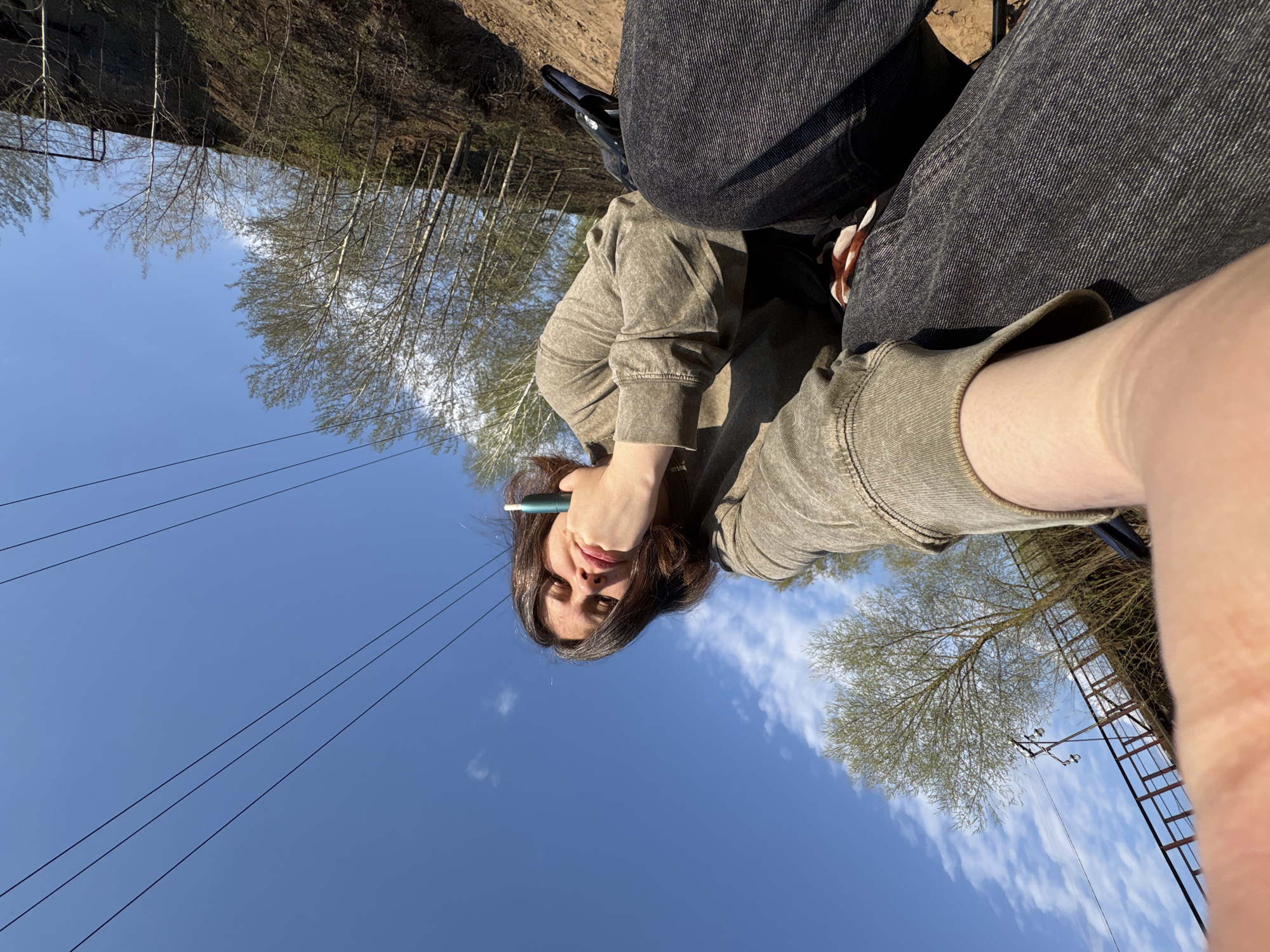 The image is taken from a low angle, looking up at a woman seated outdoors. The woman is wearing a light brown, long-sleeved sweatshirt and dark-colored jeans. She holds a green, cylindrical object to her face with her left hand. A partial view of a person's legs in dark jeans is in the background. Above the woman and in the background, the sky is blue with some clouds. There are bare trees and a utility pole with a ladder structure visible in the background. Electric wires stretch across the sky. Based on the presence of the utility pole and trees, it is likely a park or similar outdoor setting. This image was taken in the city of Chaikovsky, Russia.