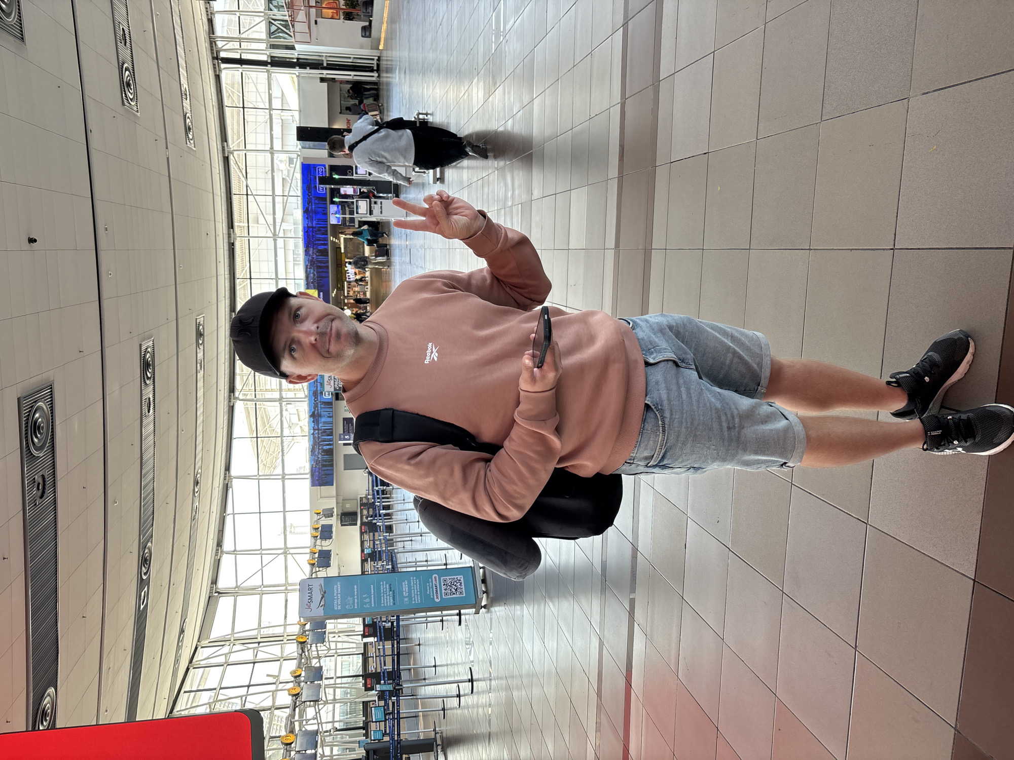 A man wearing a black baseball cap, a light pink sweatshirt with the Reebok logo, and light blue denim shorts stands in an airport terminal. He is holding a smartphone in his right hand and is making a peace sign with his left hand. He has a black backpack on his shoulders. The airport terminal has a tiled floor and a large glass ceiling that lets in natural light. In the background, there are rows of empty check-in counters, some digital signs, and other travelers. One prominent sign reads "JetSMART" and "JETSMART.COM", indicating the airline and its website. The man appears to be in good spirits. The time of day appears to be daytime due to the natural light.