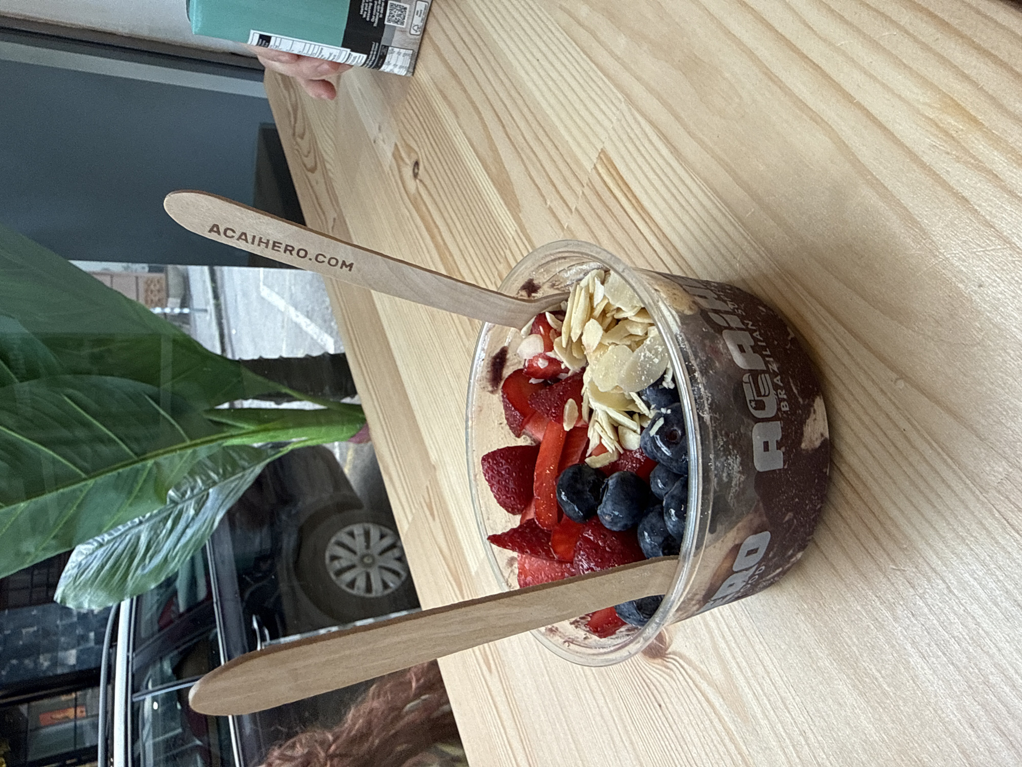 The image, oriented vertically, displays an acai bowl positioned on a light-colored wooden table with distinct grain patterns. The clear plastic bowl is filled with a deep purple acai base, generously topped with an assortment of fresh fruits and nuts. Sliced red strawberries and whole dark blueberries are prominently featured, along with a scattering of light-colored slivered almonds.
Two light-colored wooden spoons are inserted into the bowl, ready for use. Both spoons are engraved with the website "ACAIHERO.COM". The side of the plastic bowl also carries branding, with the text "ACAI HERO" and "BRAZILIAN SUPERFOOD" partially visible in dark lettering.
The scene appears to be an indoor setting, likely a cafe or restaurant in Sofia, Bulgaria, characterized by the wooden tabletop and the large window to the left. Through the window, an outdoor environment is visible, including the wheel and tire of a dark vehicle, suggesting a street or parking area. A large green plant leaf, possibly from an indoor plant, frames the left side of the image. In the upper left corner, a hand is briefly visible holding a light blue object, possibly a package. The natural lighting indicates it is daytime. The image captures a casual moment with a healthy, ready-to-eat acai bowl.