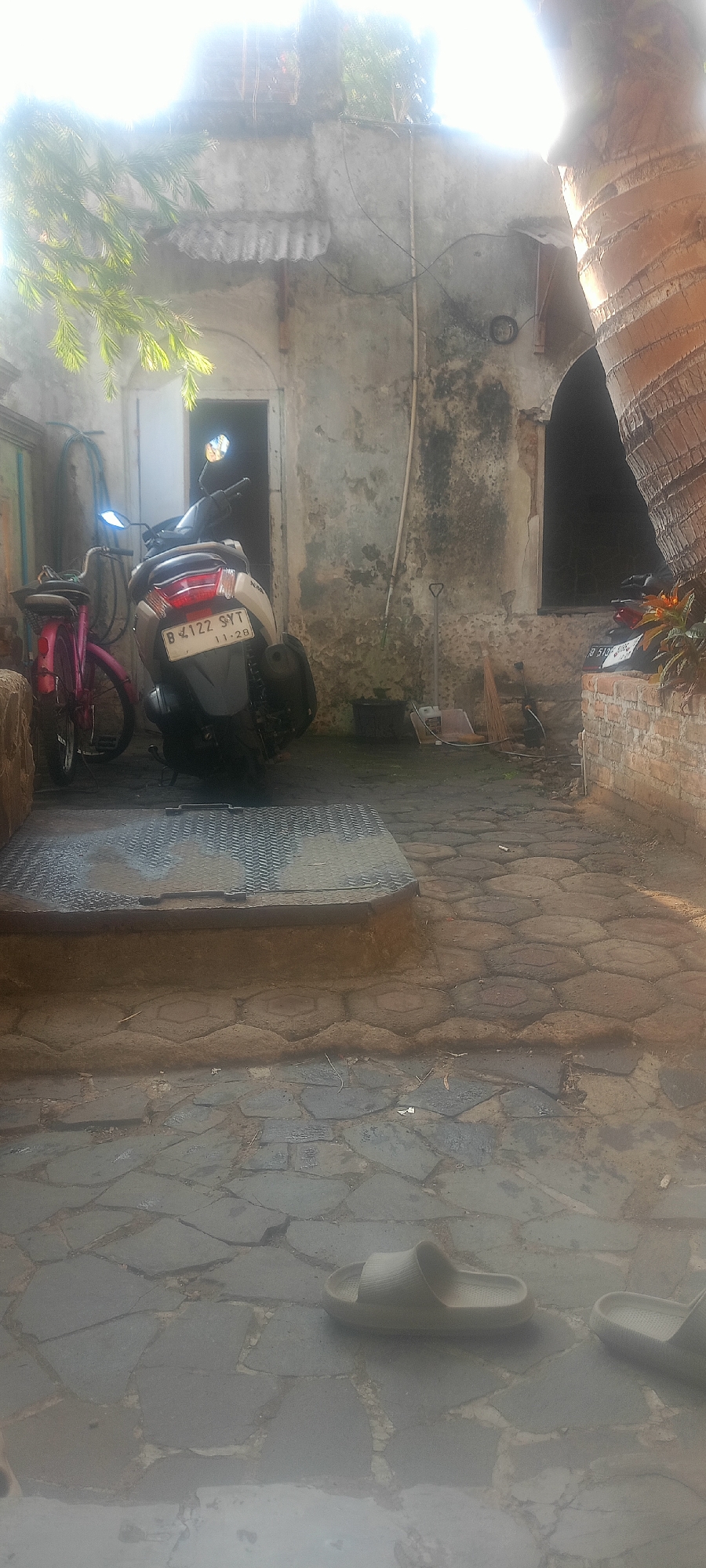 In a courtyard in Mekar Rahayu, Indonesia, a light-colored scooter and a pink bicycle are parked near a weathered, dilapidated building. The building's facade is stained and crumbling, with a dark doorway visible. The ground is paved with irregular-shaped stones and some hexagonal pavers. A metal grate covers a portion of the ground in the foreground. In the lower portion of the image, two light-colored slip-on sandals are lying on the stone pavement. A large palm tree trunk is visible on the right side of the frame, and foliage from another tree hangs from the upper left. The lighting suggests it is daytime, with bright sunlight creating some glare.