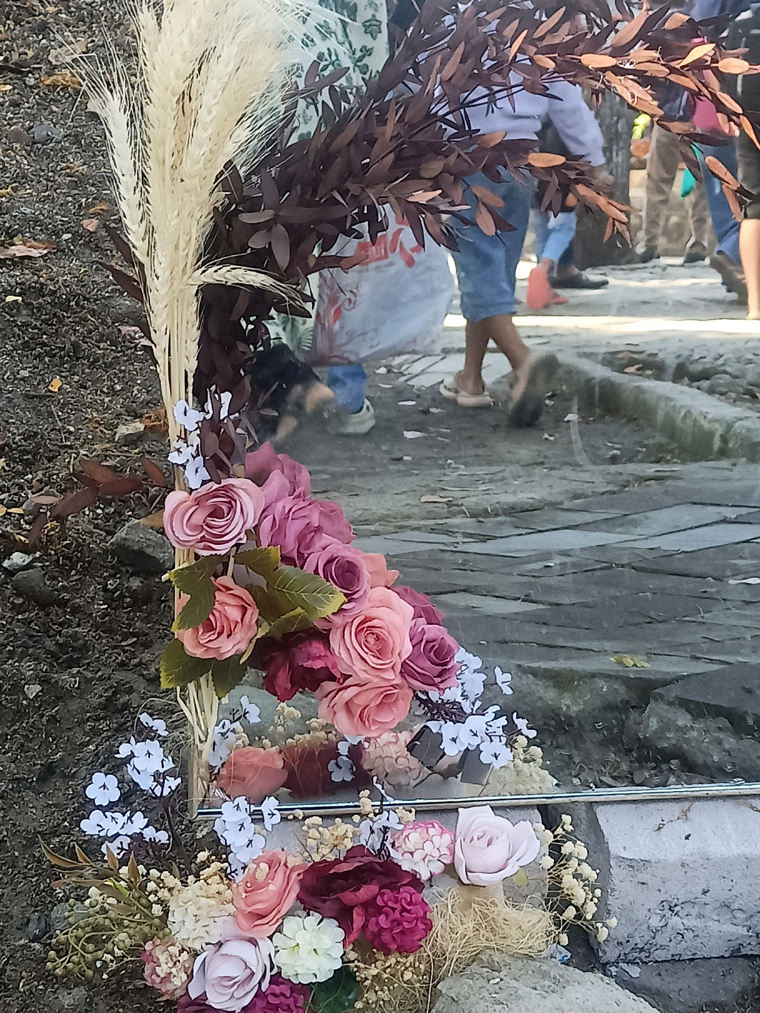 The image depicts an outdoor scene with a focus on floral arrangements in the foreground. A bouquet of artificial flowers, primarily in shades of pink and burgundy, is arranged near the bottom of the frame. These flowers are interspersed with dried grasses and small white floral accents. Behind this, a taller arrangement features dried wheat stalks and dark brown leaves.
The background is blurred, but it appears to be a public area with people walking on a cobblestone or paved walkway. Some individuals are wearing casual clothing, including denim shorts and t-shirts. There is a dog visible in the middle ground, looking towards the flowers. The environment appears somewhat unkempt, with loose soil and gravel visible around the edges. The scene is illuminated by natural daylight. The provided context indicates this location is in Karungan, Indonesia.